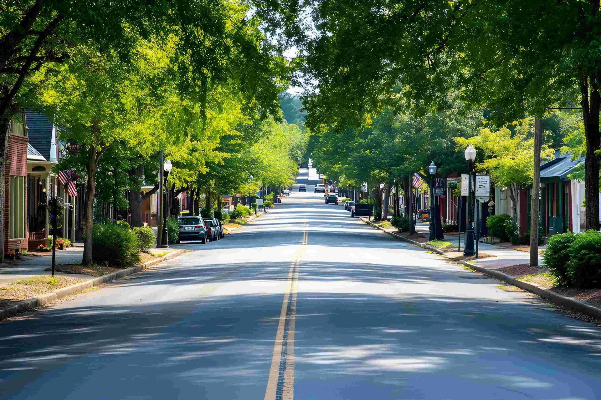 A sunny, tree-lined downtown street with old storefronts, street lamps, parked cars, and American flags