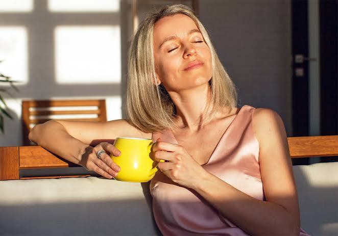 Woman in warm golden sunlight holding a mug, eyes closed in a moment of peace