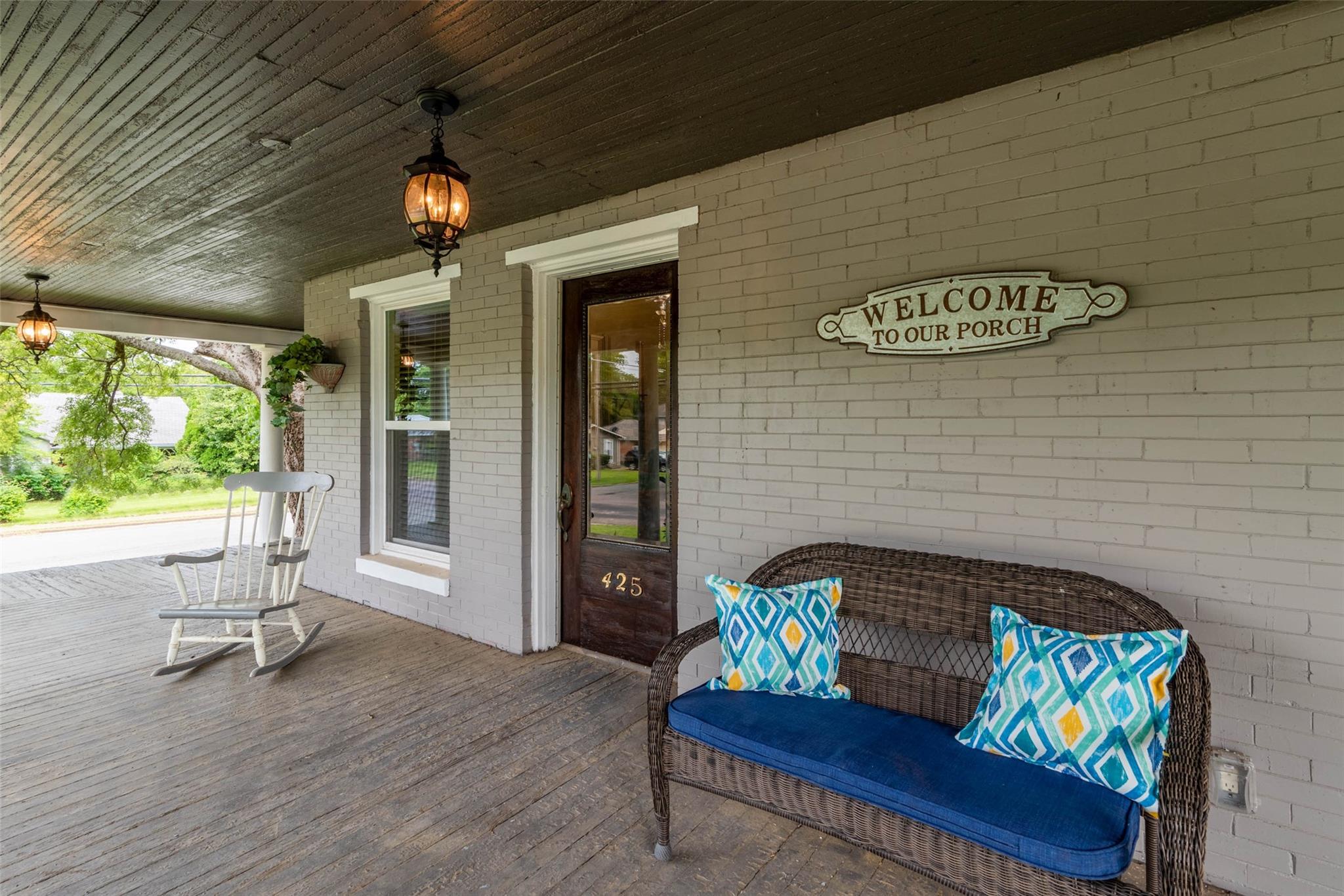 Warm front porch with a painted brick wall, a wooden WELCOME TO OUR PORCH sign, a white rocking chair, and a wicker bench with a blue cushion and patterned pillows