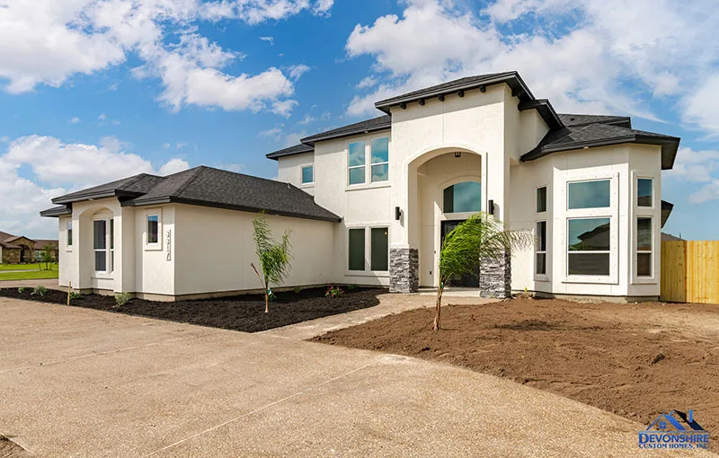 a newly constructed two-story modern single-family home with off-white stucco walls, a dark grey shingled roof, stone accents around the entrance, a concrete driveway, and a freshly landscaped yard with young palm trees under a bright blue sky