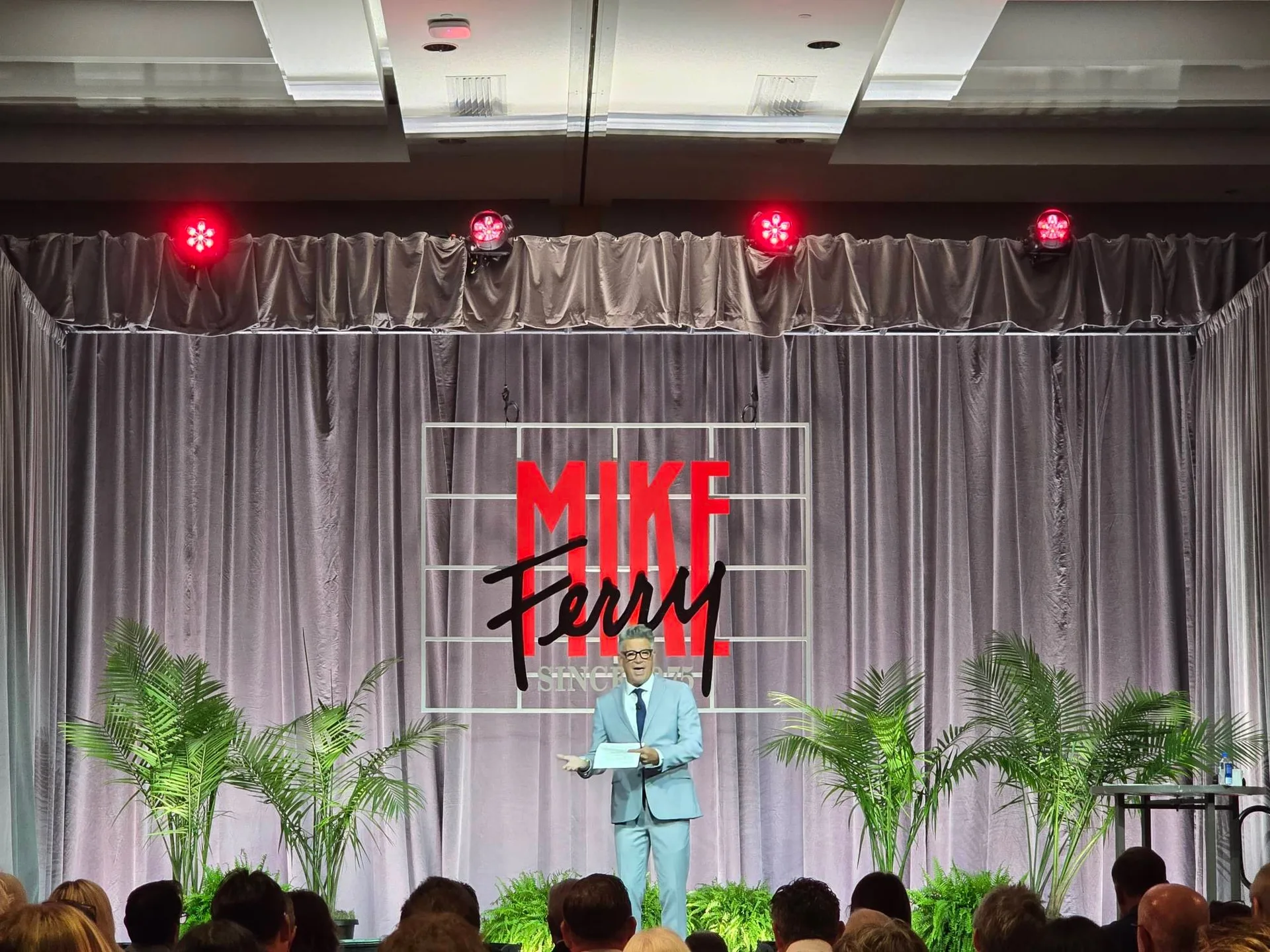Ron Cronin speaking to the audience on stage in a light blue suit and glasses at the Mike Ferry 2026 Superstar Retreat at The Diplomat Beach Resort in Hollywood, Florida, with the MIKE FERRY backdrop, red stage lights, palm plants, and the audience seated in front
