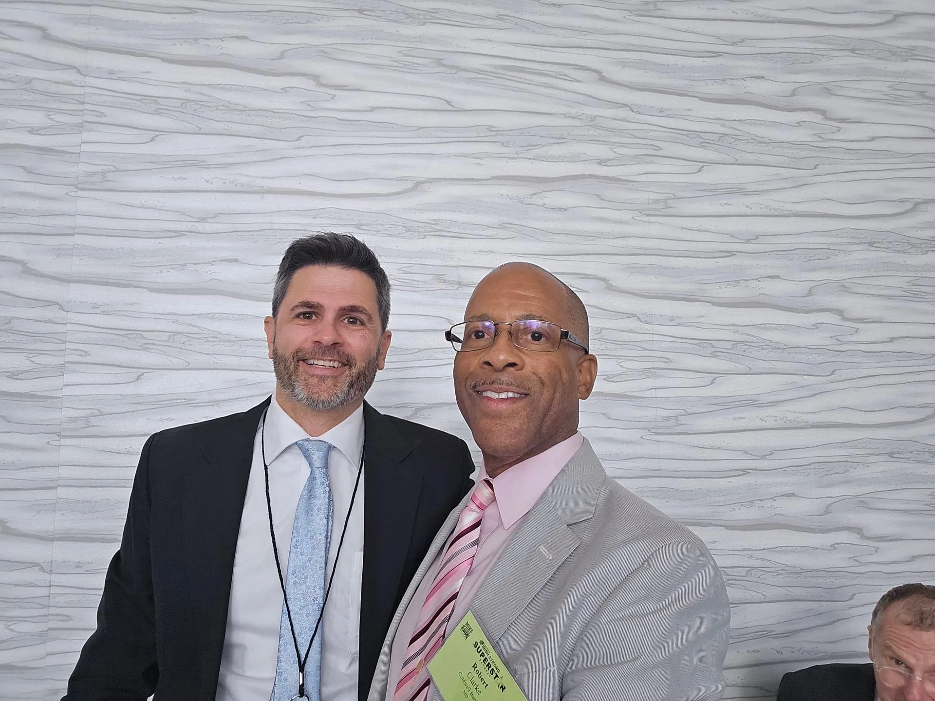 Robert Clarke poses with another conference attendee, both in suits and ties with conference badges, standing against a light wood-textured wall at the Mike Ferry 2026 Superstar Retreat