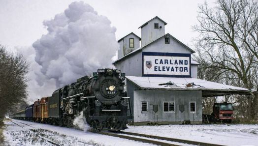 A restored black steam locomotive with golden trim beside a red-brick 1940s roundhouse and repair shop in Owosso, Michigan on a crisp autumn afternoon