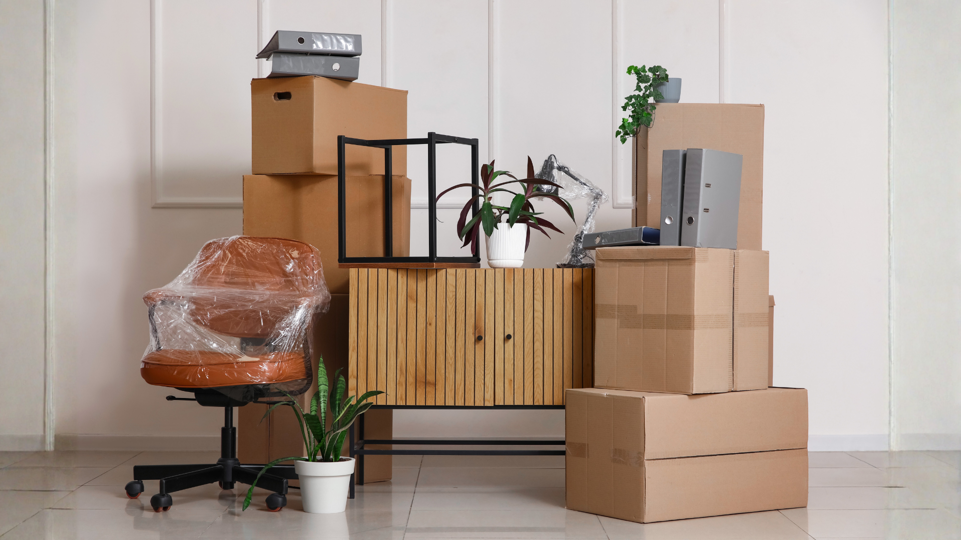 Stacked cardboard moving boxes, a wrapped office chair, and plants during a home relocation
