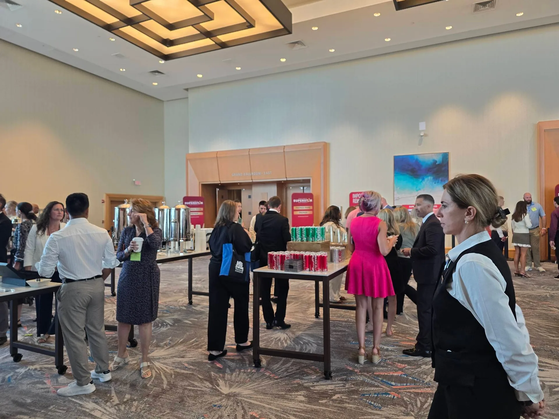 Attendees in business attire mingling around a refreshment table with red soda cans and coffee urns during a break at the Mike Ferry 2026 Superstar Retreat at The Diplomat Beach Resort in Hollywood, Florida, with red event banners visible in the background