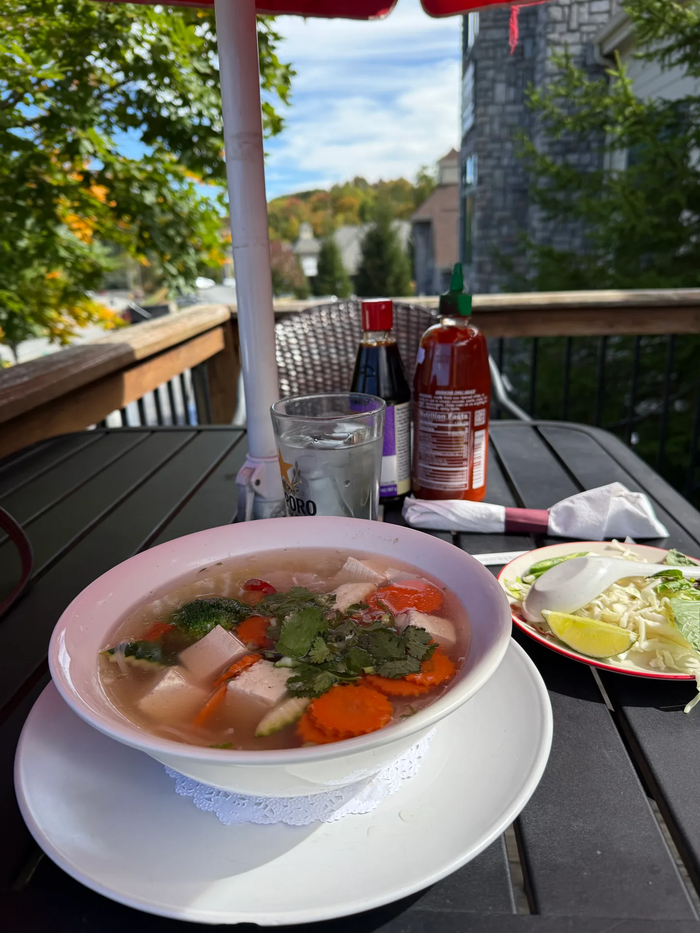 A steaming bowl of vegetarian pho with tofu, carrots, cilantro, and bean sprouts on the patio at Luna Thai & Sushi in Banner Elk, with lime and sriracha on the table