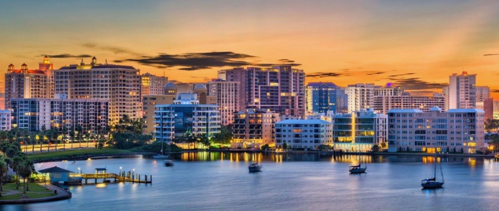 Sarasota waterfront at sunset with boats on the bay, palm trees, and the city skyline glowing in warm golden-hour light