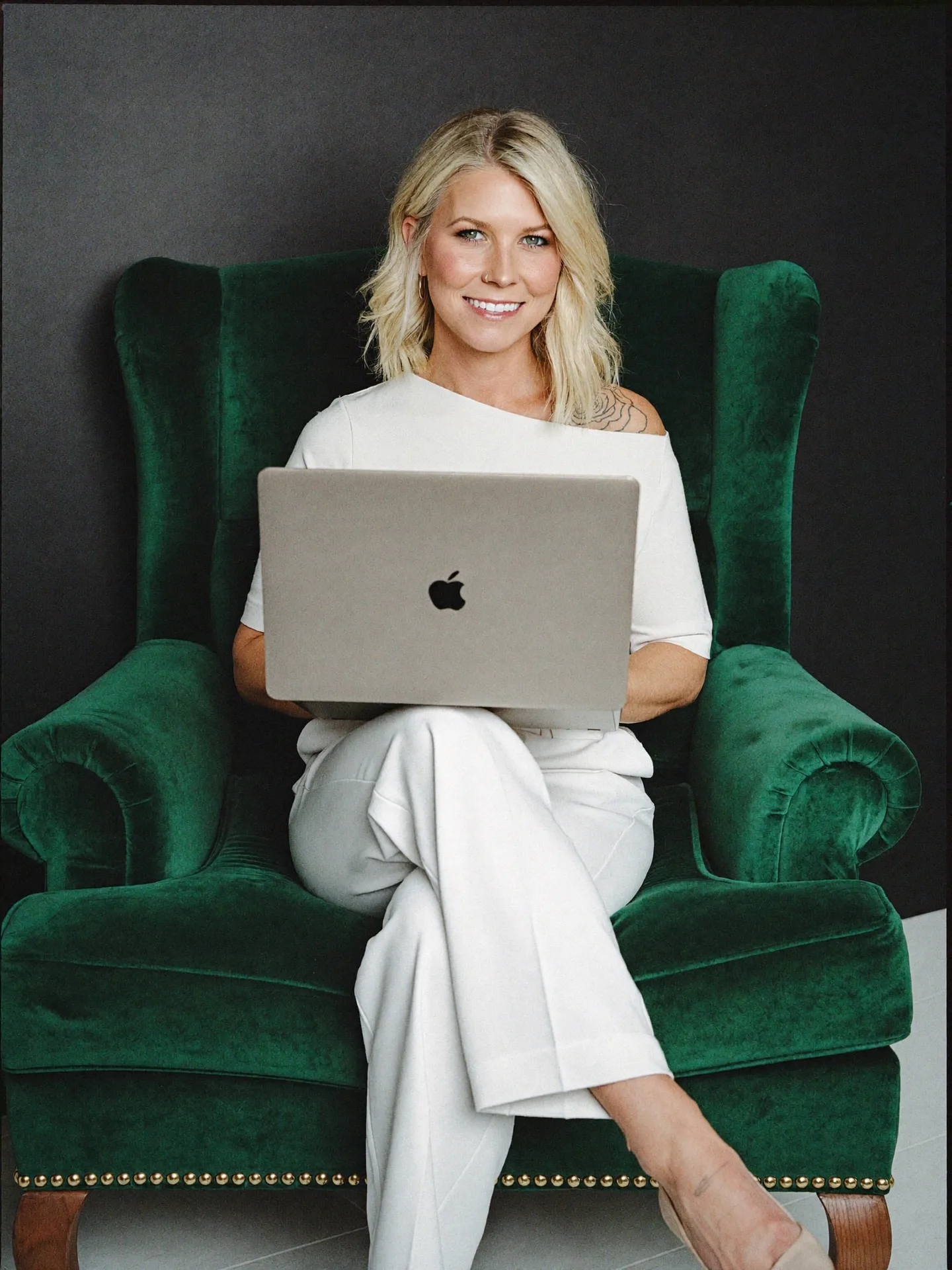 Nicole Elizabeth Yaeger relaxing in a green velvet wingback chair with a laptop, smiling in her element