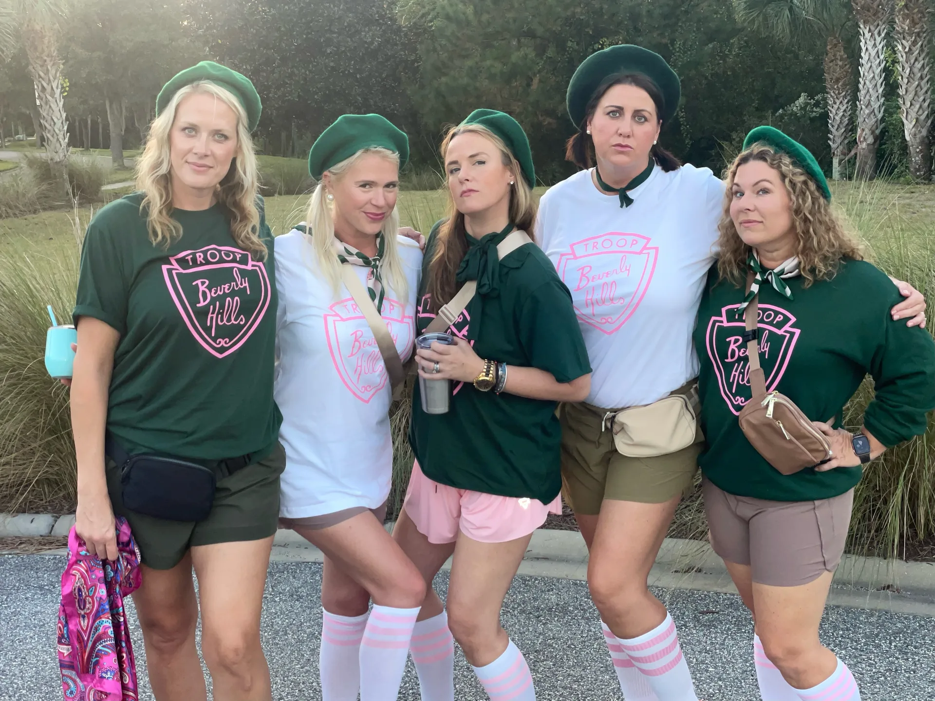 Five women in matching Troop Beverly Hills costumes with green berets and shields, posing outdoors at a Halloween celebration