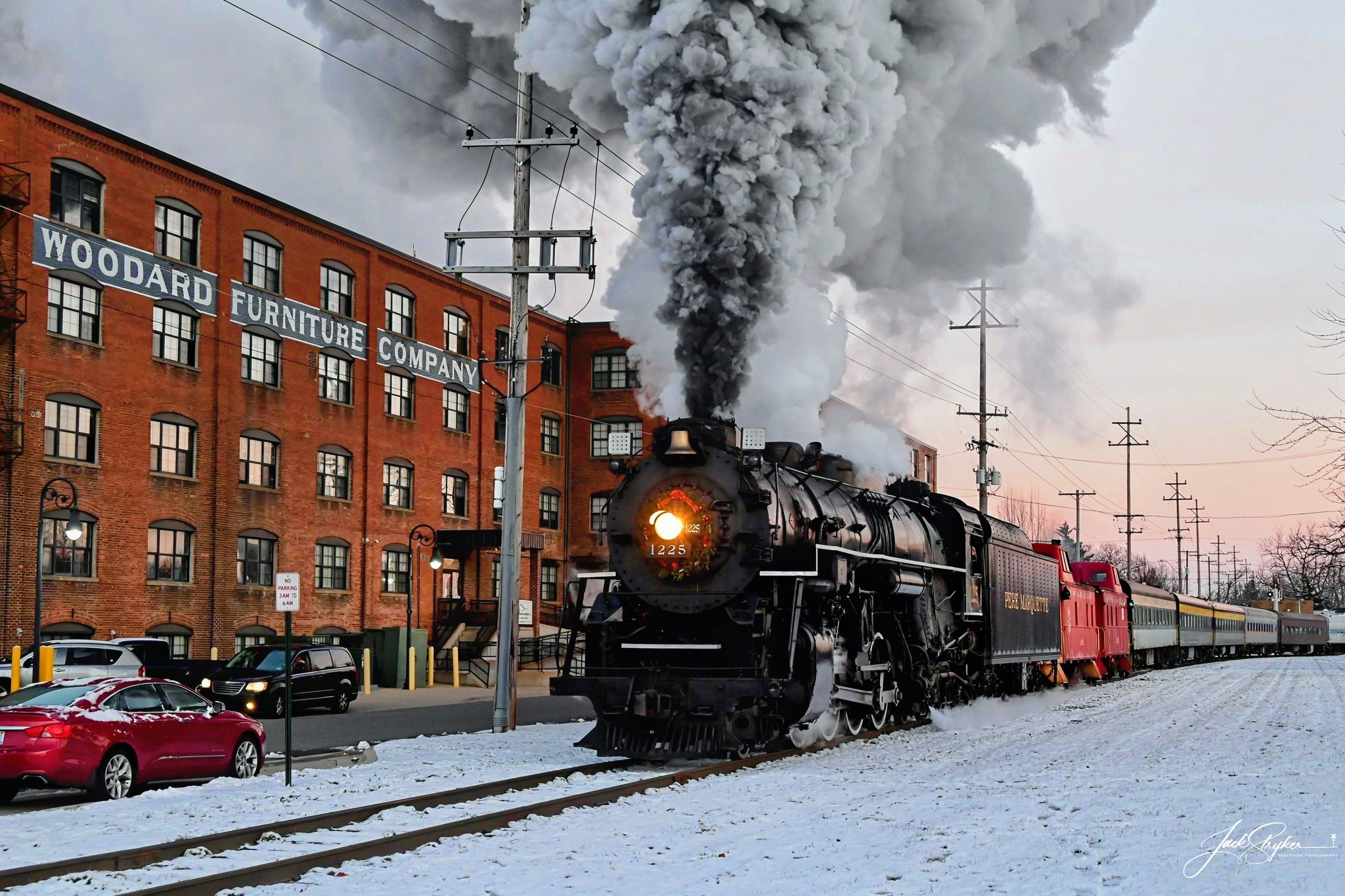 A classic black steam locomotive with a brass bell and golden trim standing at a small-town Michigan red-brick railroad station, steam curling from the smokestack on a sunny morning