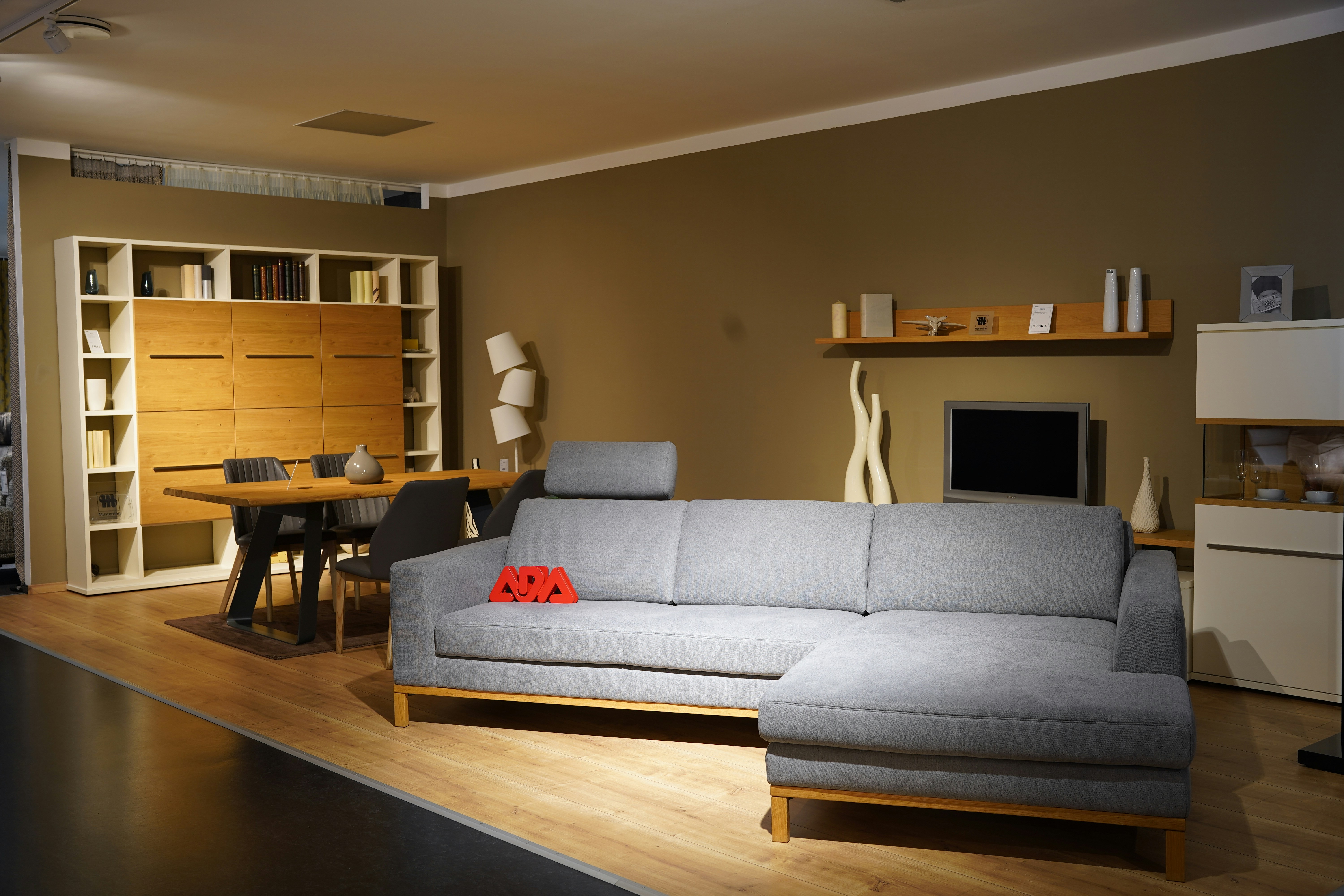 Modern minimalist showroom interior with grey L-shaped sectional sofa, dining area, warm ambient lighting, and clean styling