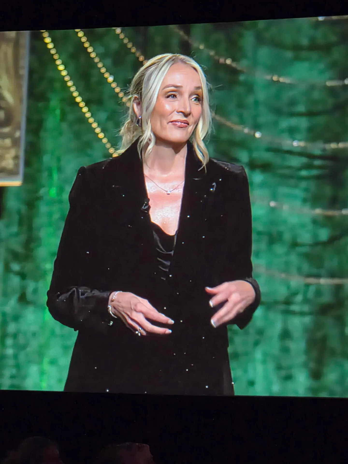 An attendee in a black blazer stands in front of a green backdrop with string lights at the Better Homes and Gardens Real Estate retreat