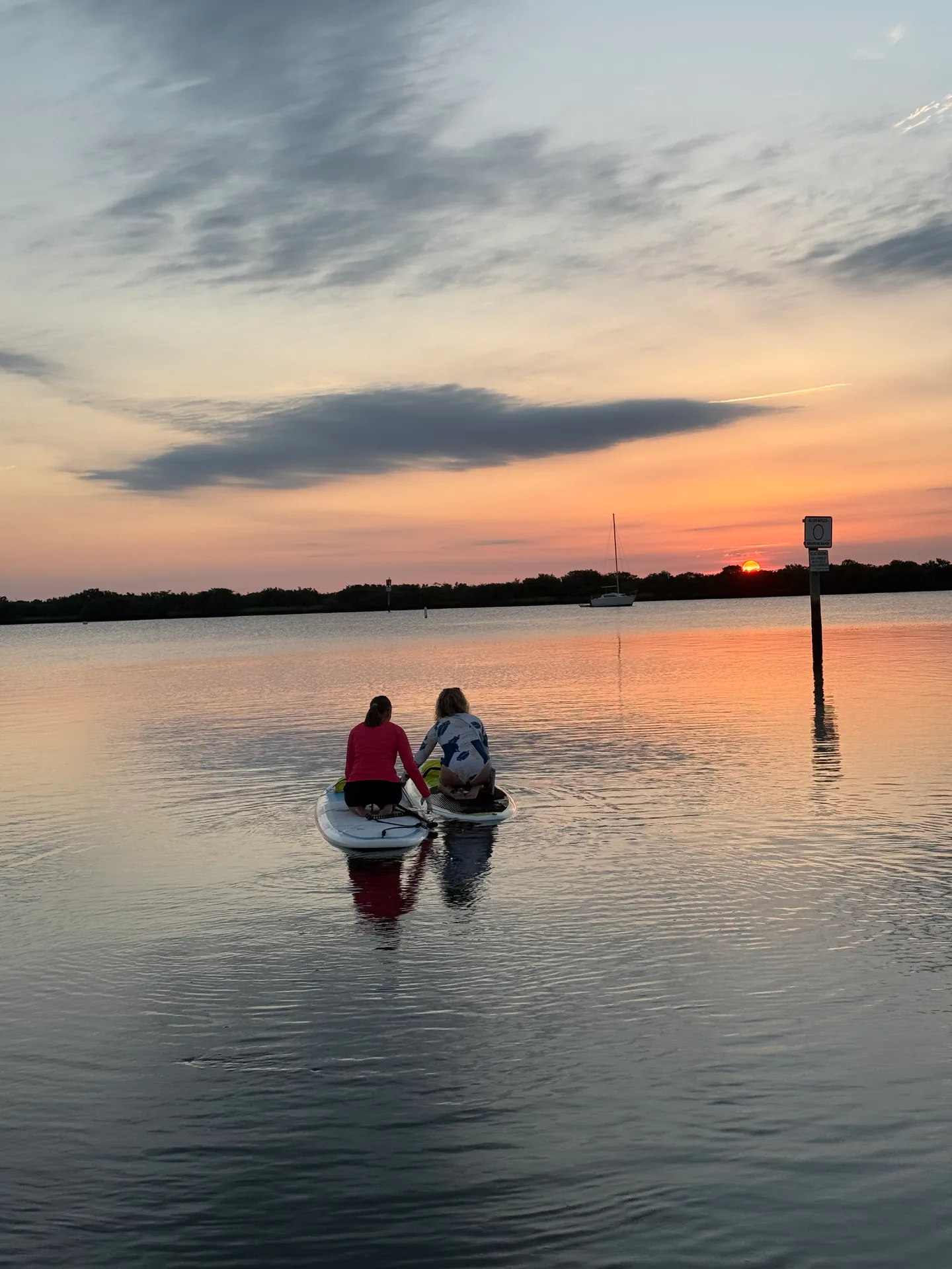 Two people stand-up paddleboarding at sunset on calm water with a shoreline and a small sailboat in the distance
