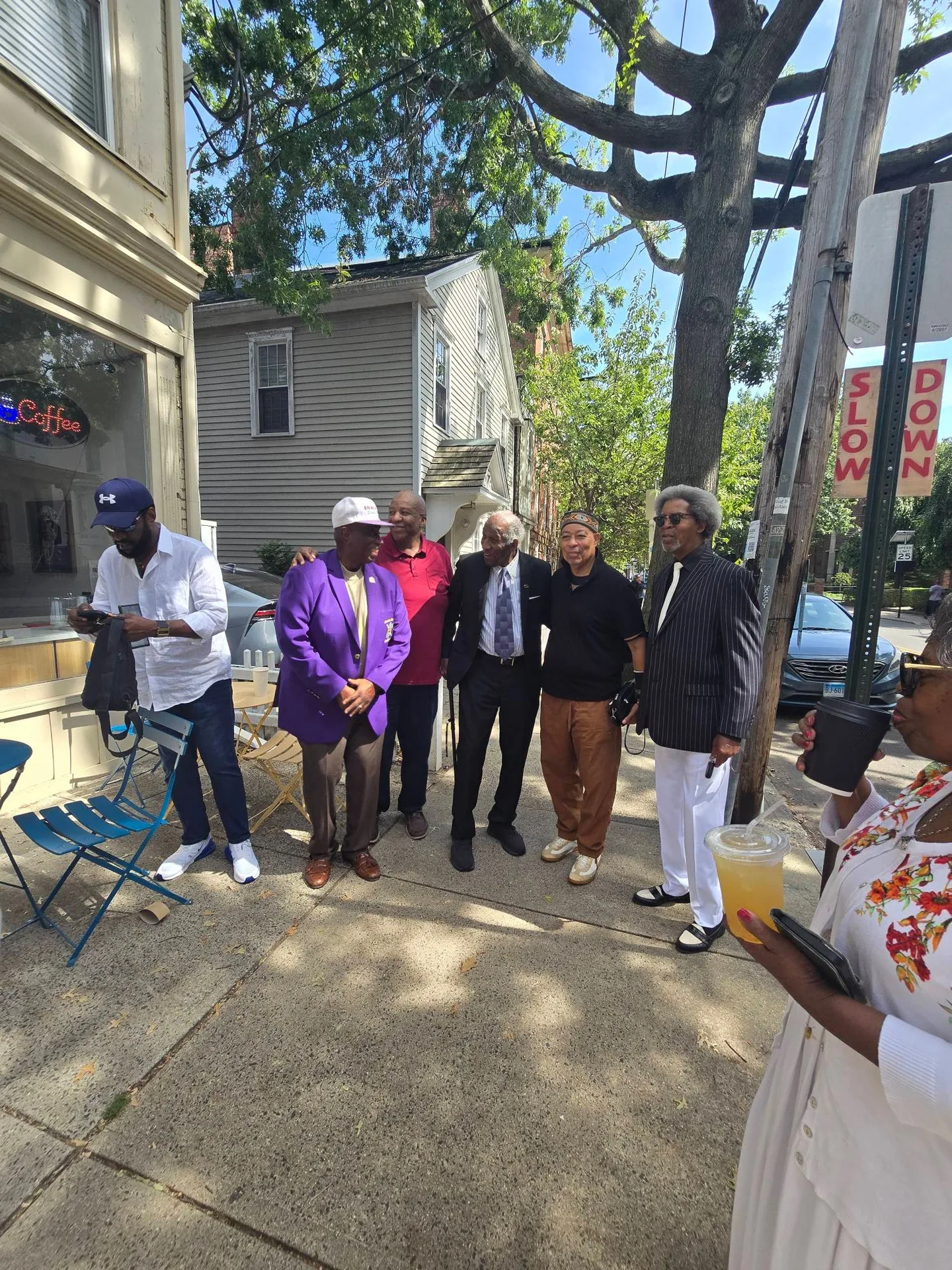 Six men gathered outdoors on the sidewalk in front of the cafe, with the Coffee neon sign visible in the window