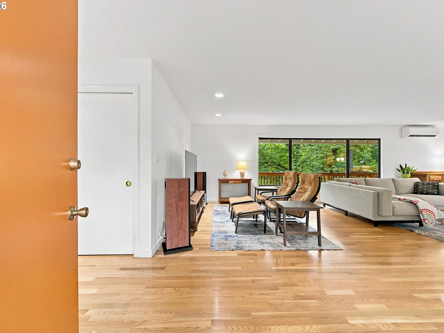 Open-concept living area at 1111 SW Coronado St with hardwood floors, forest views through large windows, and the mini-split wall unit visible in the background