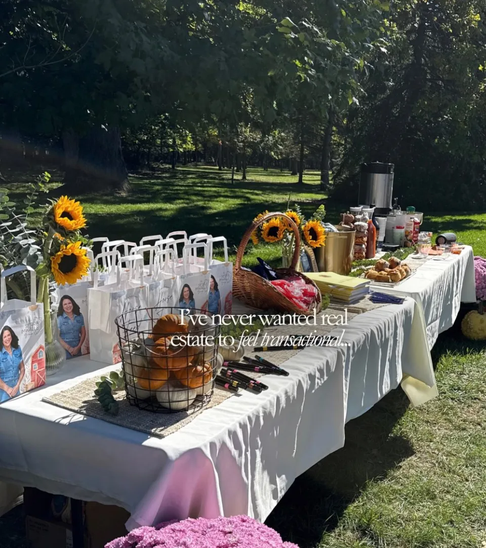 Sunny fall client appreciation event hosted by Sierra Imwalle: a long white-clothed table with white gift bags, sunflowers, small pumpkins, and snacks in a grassy area with trees