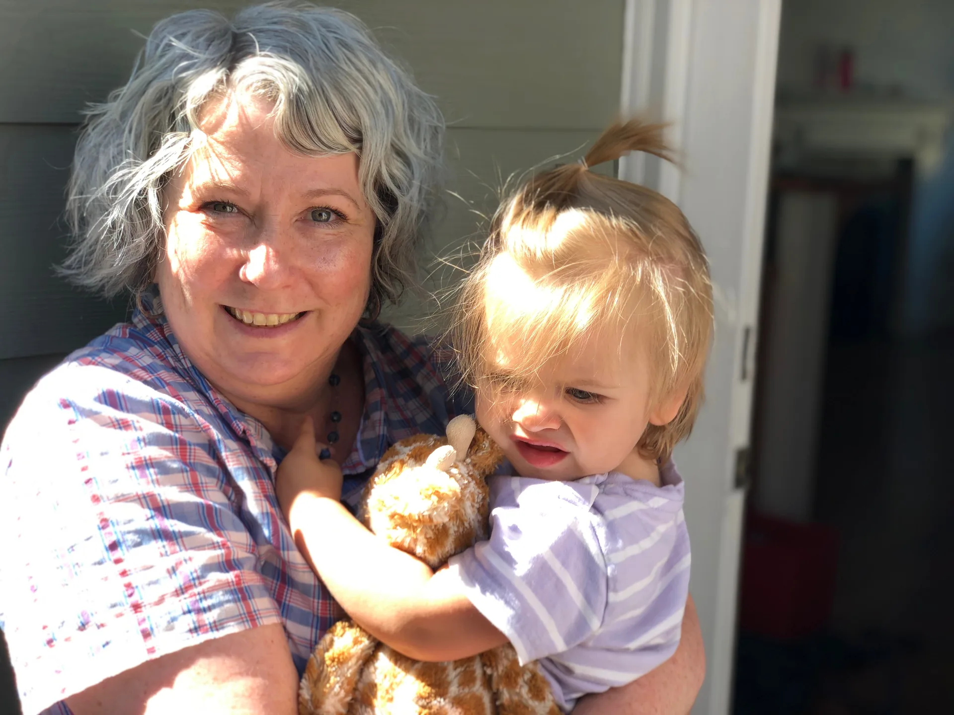A woman holds a young child with a stuffed giraffe in a warm outdoor setting