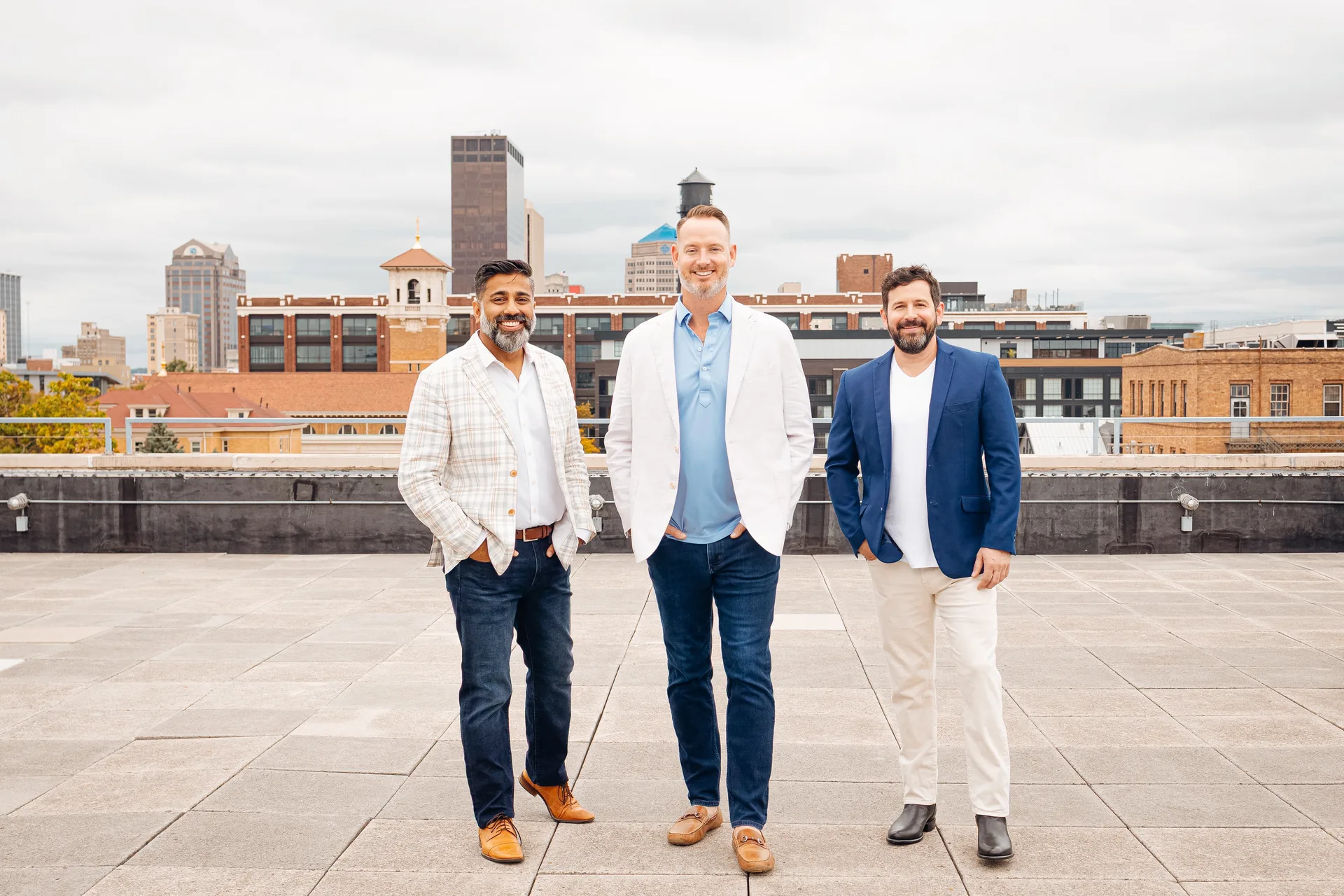 Glasshouse Realty brokers Mo Zahedi, Evan Kloth, and Kunal Patel on a rooftop in light blazers with a city skyline in the background