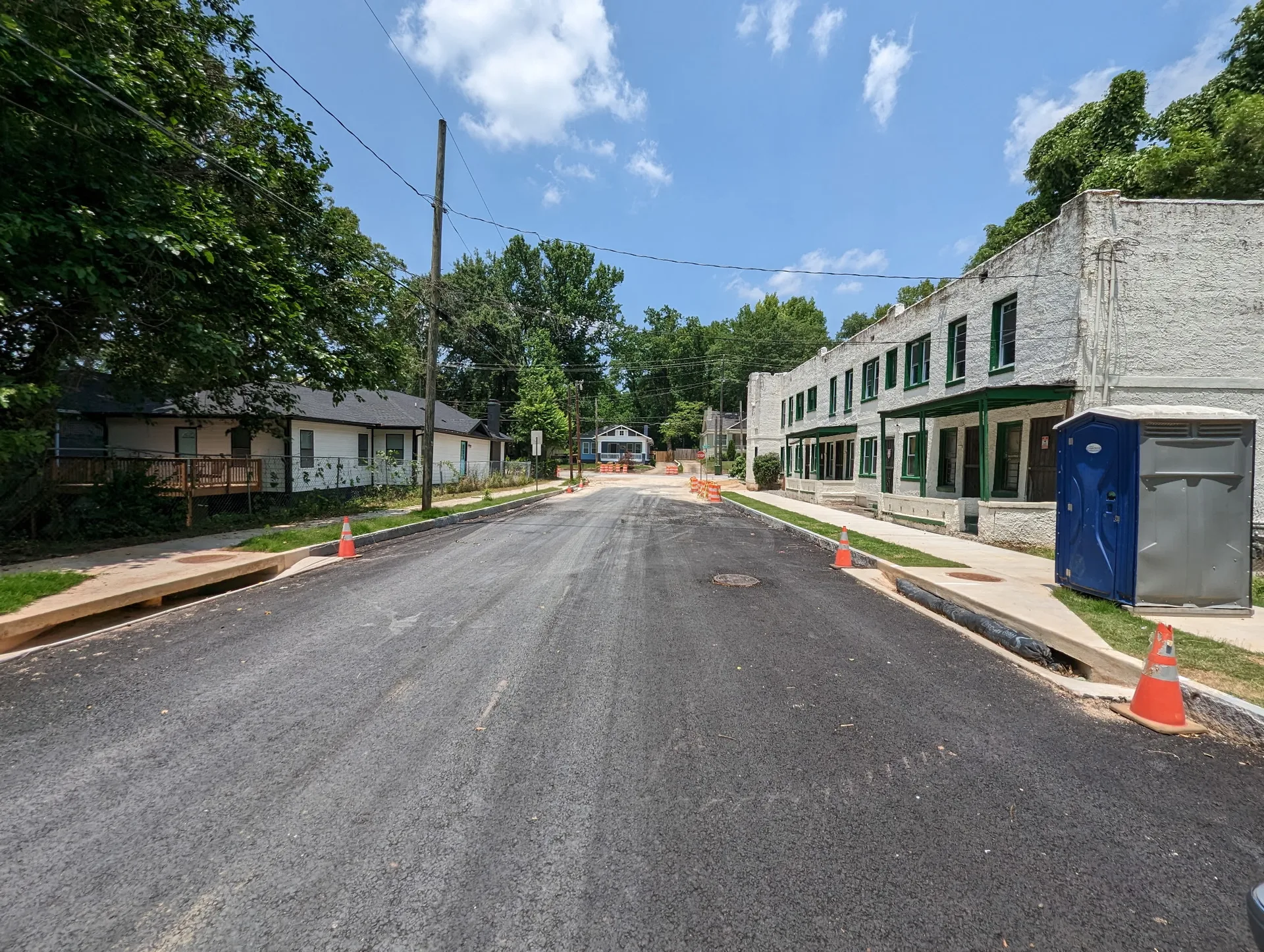 A residential street scene on a sunny day with a white multi-unit building with green-trimmed windows and doors, fresh asphalt, and mature trees
