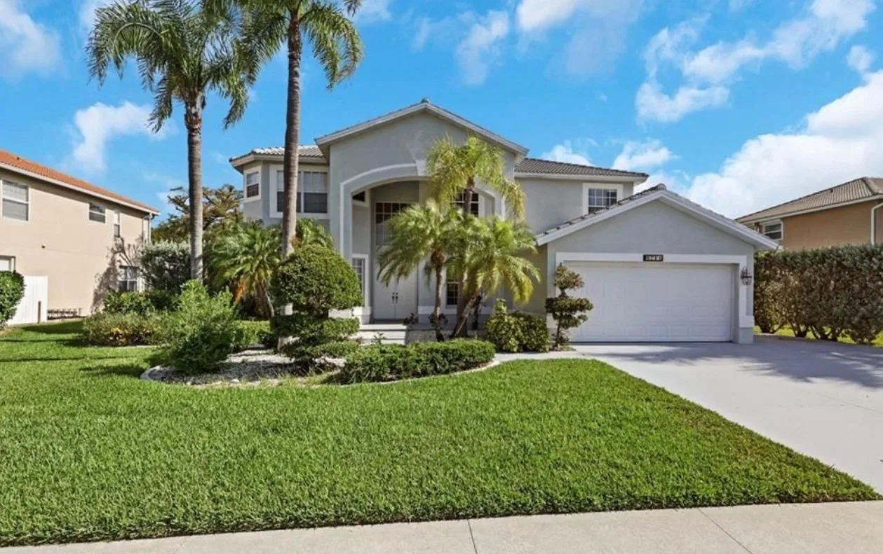 Front exterior of 8711 Kilkenny Ct, a light grey stucco home with tiled roof, arched entryway, two-car garage, and palm trees in Cross Creek Estates, Fort Myers, Florida