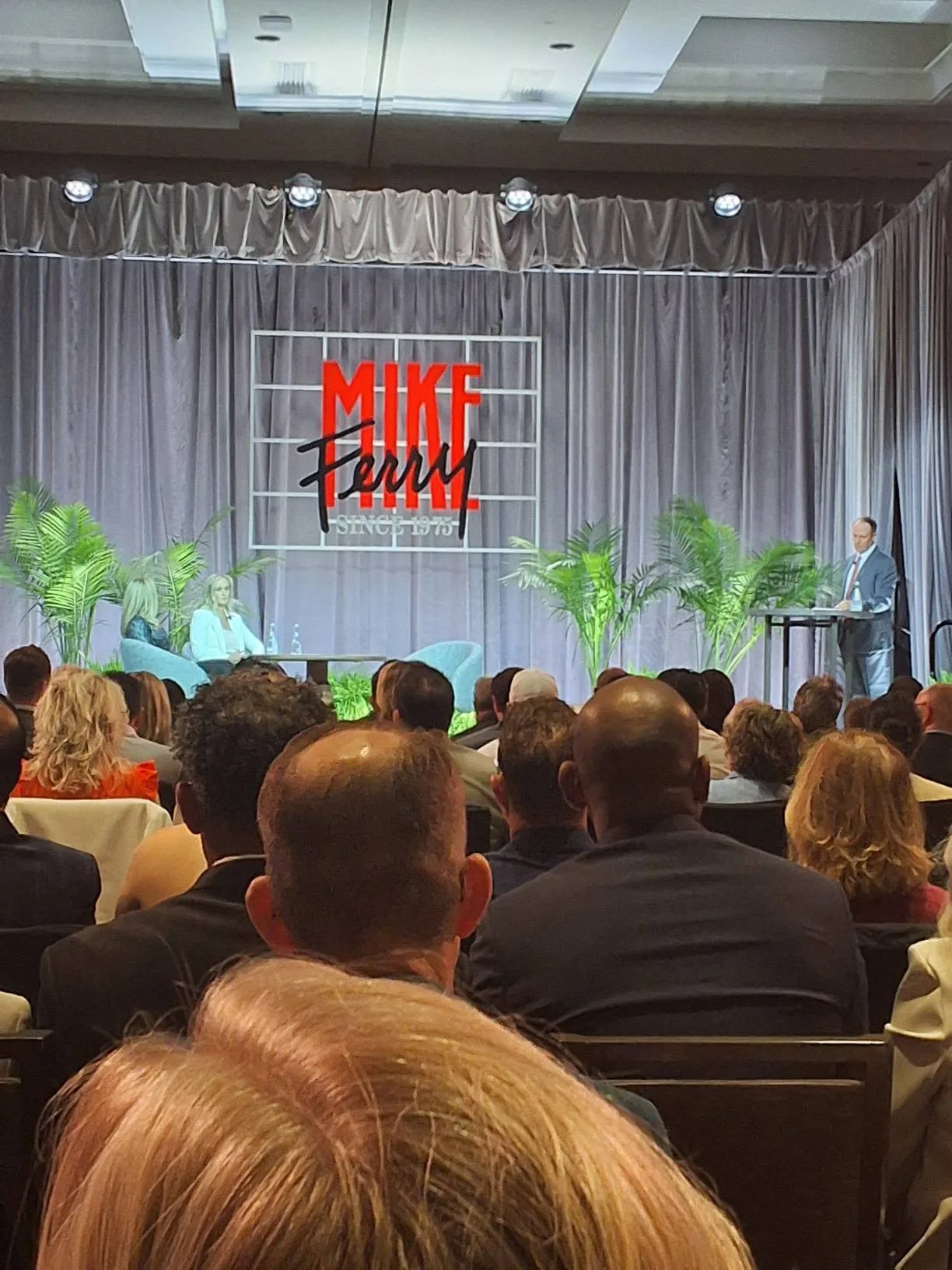 A speaker at the podium during a live on-stage session at the Mike Ferry 2026 Superstar Retreat at The Diplomat Beach Resort in Hollywood, Florida, with two guests seated in blue armchairs, palm plants, the MIKE FERRY SINCE 1975 backdrop, and the audience seated in front