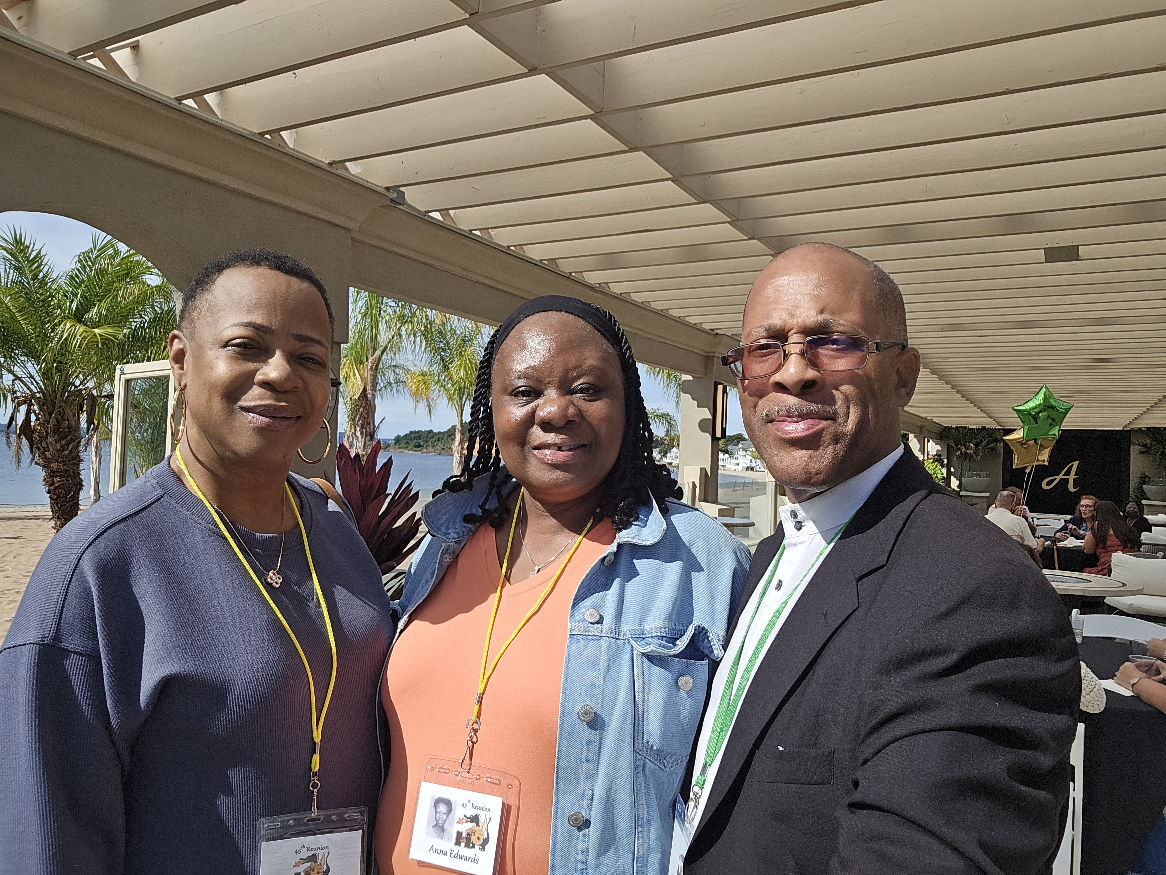 Anna in an orange top with a denim jacket at the center, Kwan in a dark grey top on the left, and Robert in a black suit jacket over a white shirt on the right, all wearing reunion badges and posing under a pergola on the waterside patio at the Hamden High Class of 1981 45th reunion.