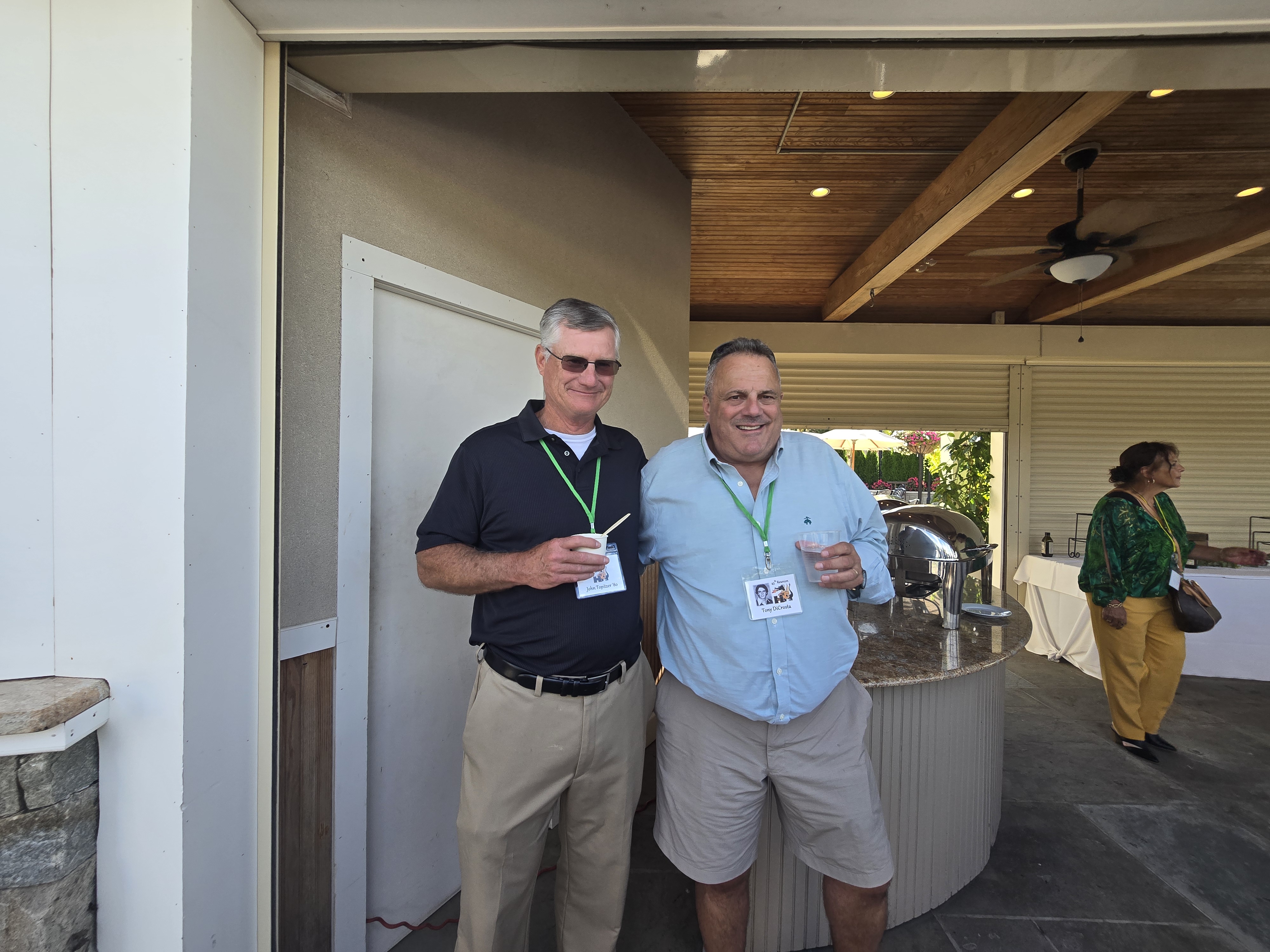 Two men smiling at the Hamden High Class of 1981 45th reunion, one in a black polo and one in a light blue button-down, both wearing green lanyards with name tags near the serving area.