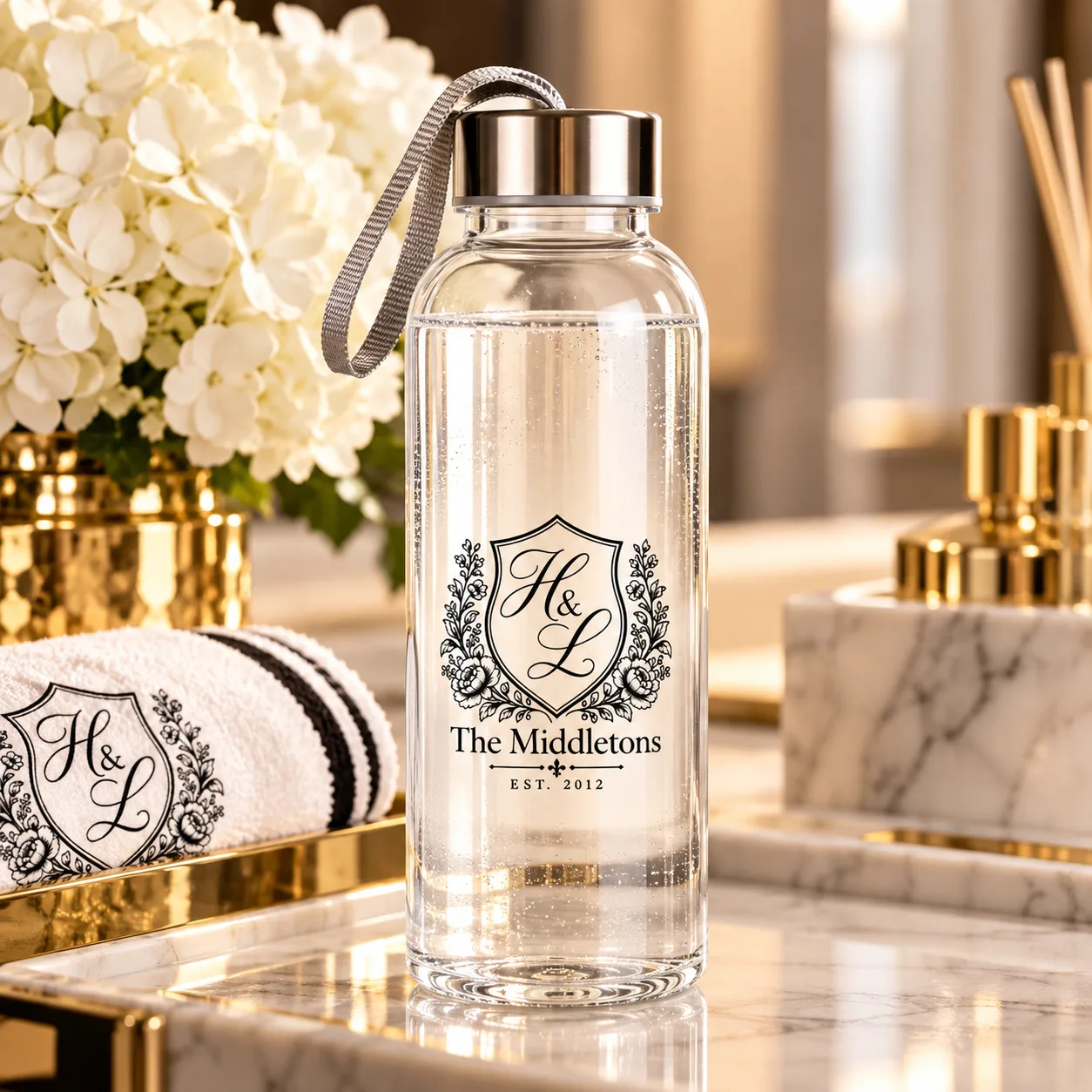 The House Merch Co. personalized glass water bottle: clear glass, silver screw cap, grey strap, and a black crest monogram reading "The Middletons, EST. 2012" inside a floral wreath, styled on a marble vanity with a monogrammed towel and white hydrangeas