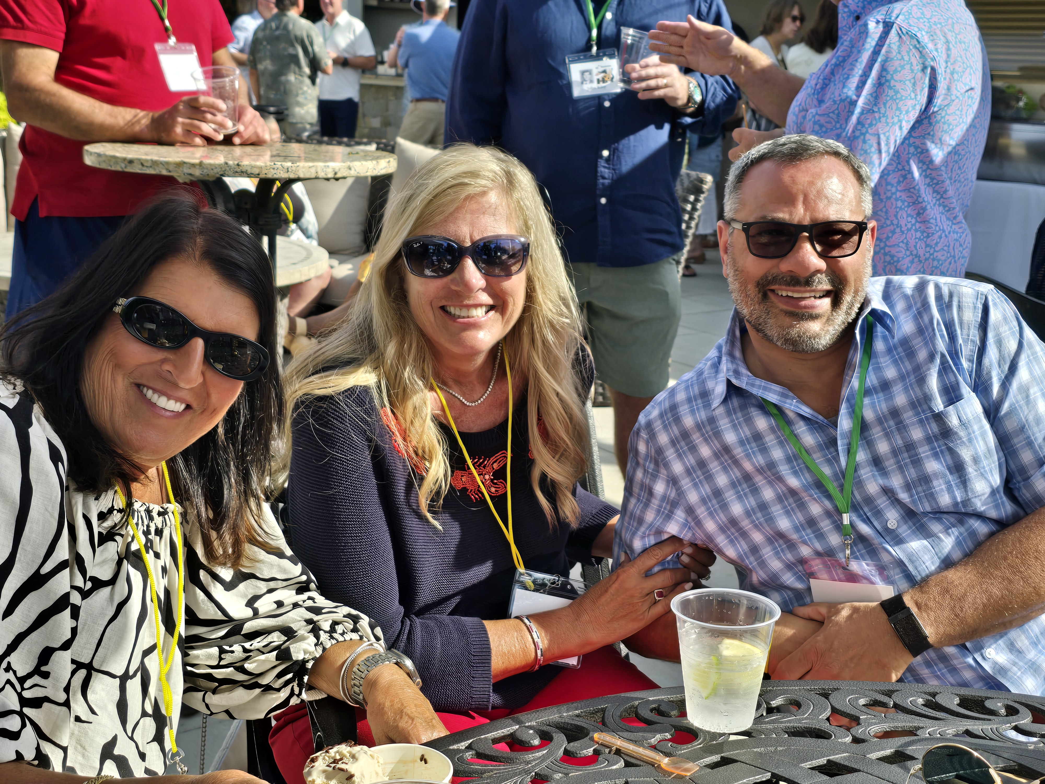 Anthony in a light blue plaid shirt, Samantha in a dark sweater in the center, and Roxy in a black and white patterned top, wearing event badges on the sunny patio at the Hamden High Class of 1981 45th reunion.