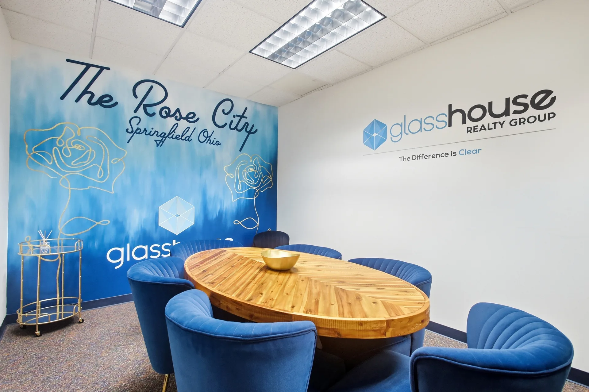 The Glasshouse Realty Springfield office conference room, with an oval wooden table and blue chairs beneath The Rose City Springfield Ohio wall art and the Glasshouse Realty Group logo