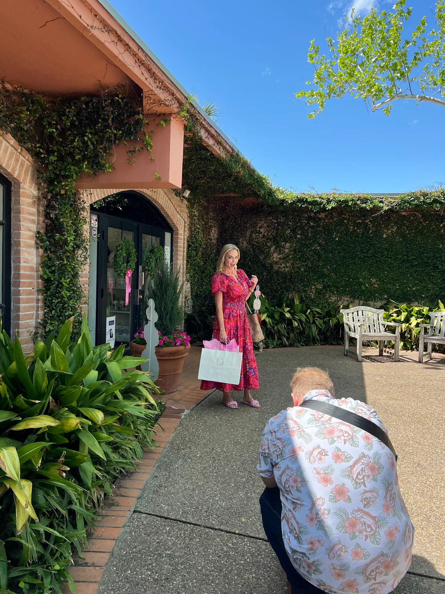 Emily Chemacki holding shopping bags in a bright pink floral-patterned dress in a sunny courtyard