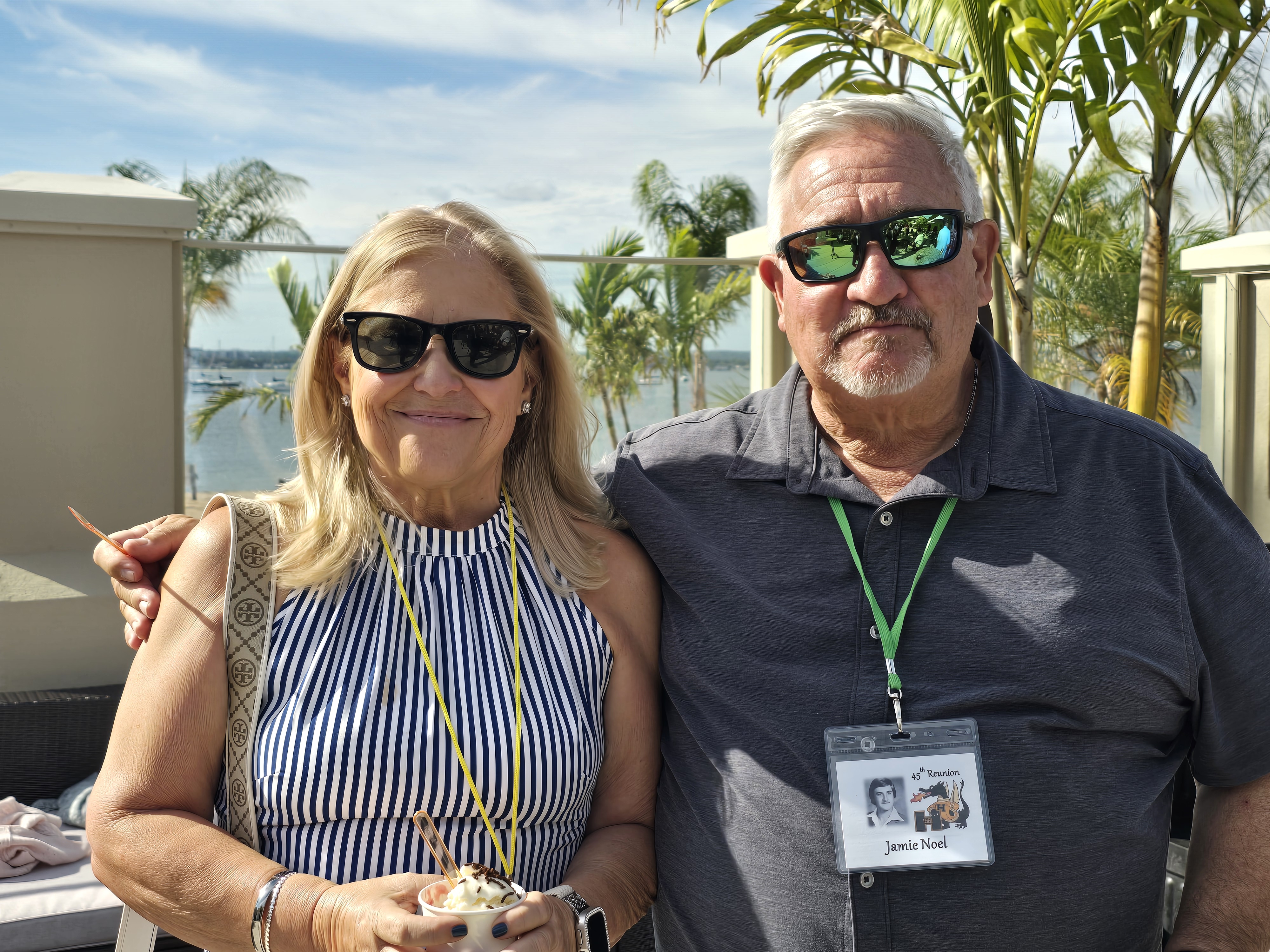 Jim with gray hair in a gray polo shirt and Vicki with blonde hair in a blue and white striped top, both wearing sunglasses and reunion badges, overlooking the water at the Hamden High Class of 1981 45th reunion.