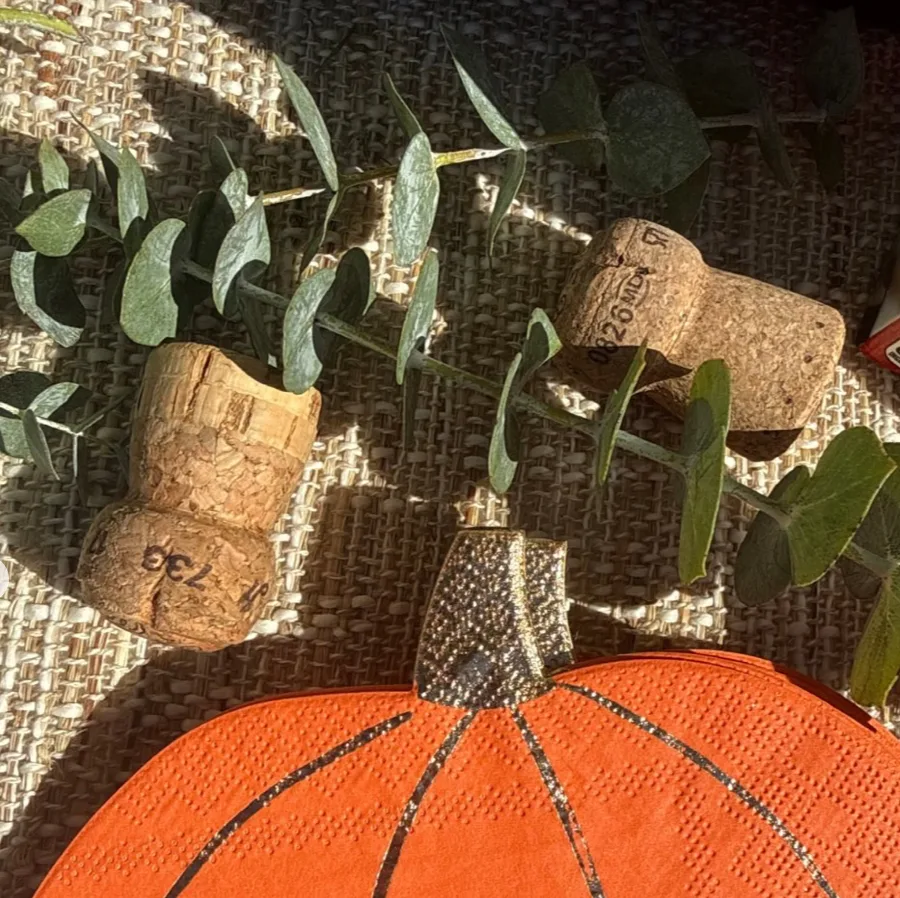 Close-up of pumpkin-shaped orange napkins with a gold stem, champagne corks, and eucalyptus on a woven surface