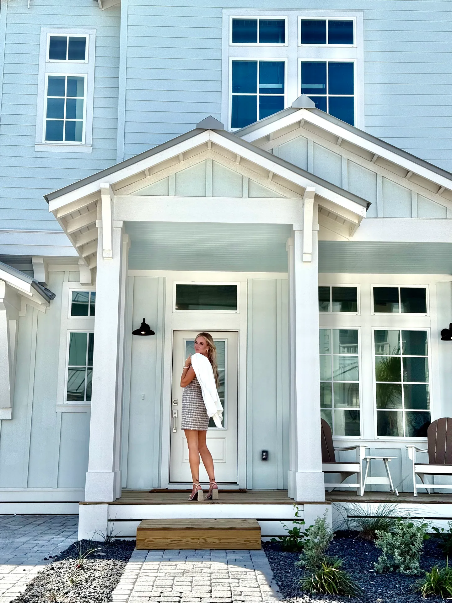 A young woman standing on the porch of a light-blue coastal-style house, looking back toward the camera with a white jacket over her shoulder