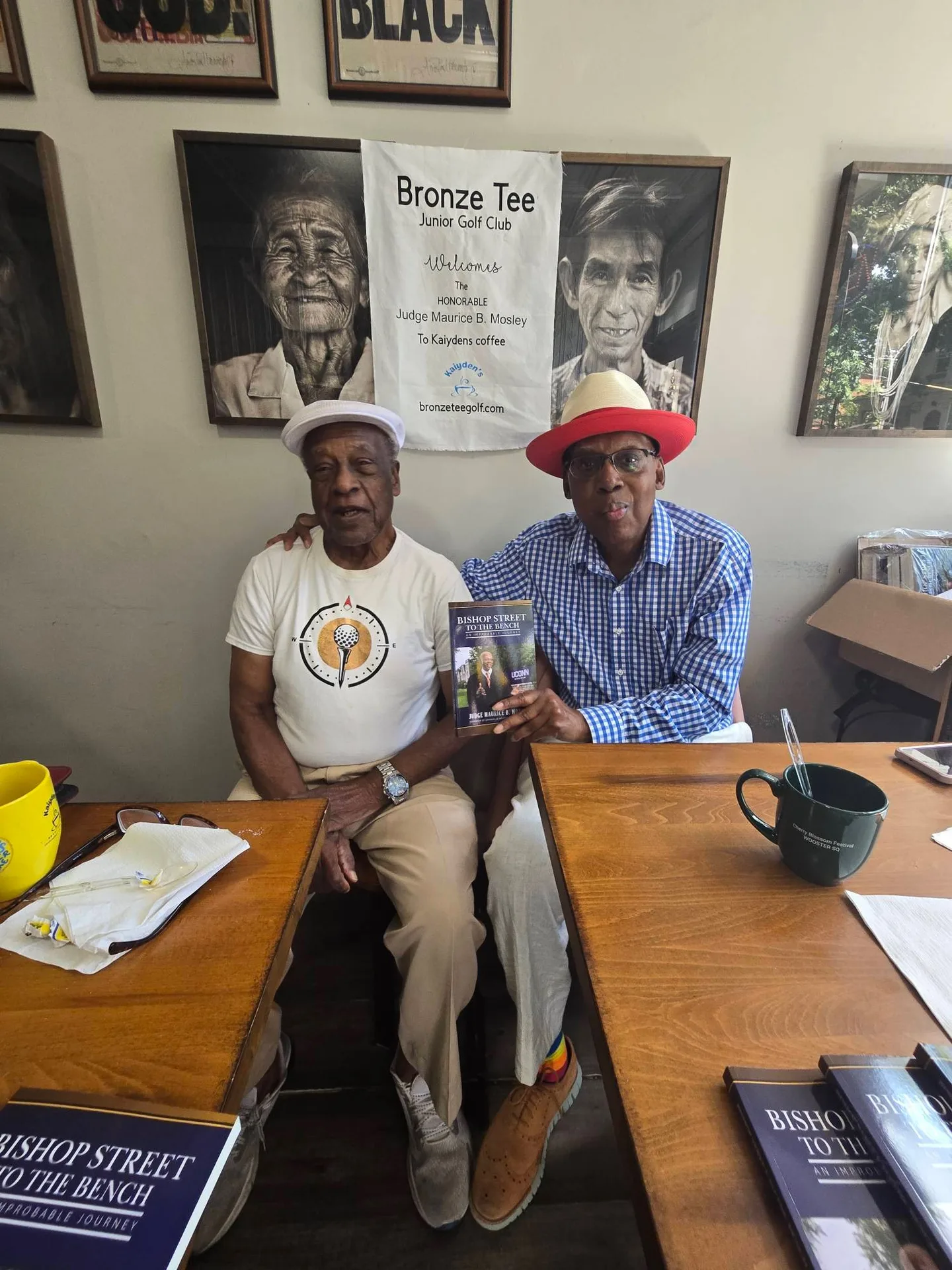 Judge Mosley in a fedora at the signing table holding a copy of Bishop Street to the Bench, with the Bronze Tee Junior Golf Club welcome banner behind him