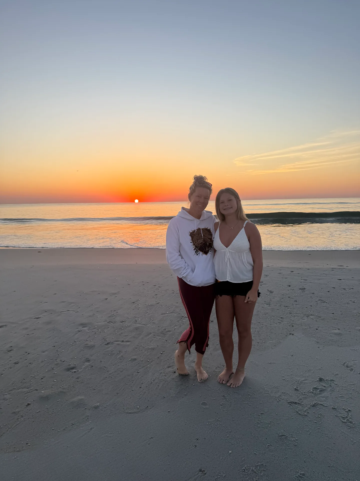 Two women standing barefoot on the beach at sunrise with calm water behind them