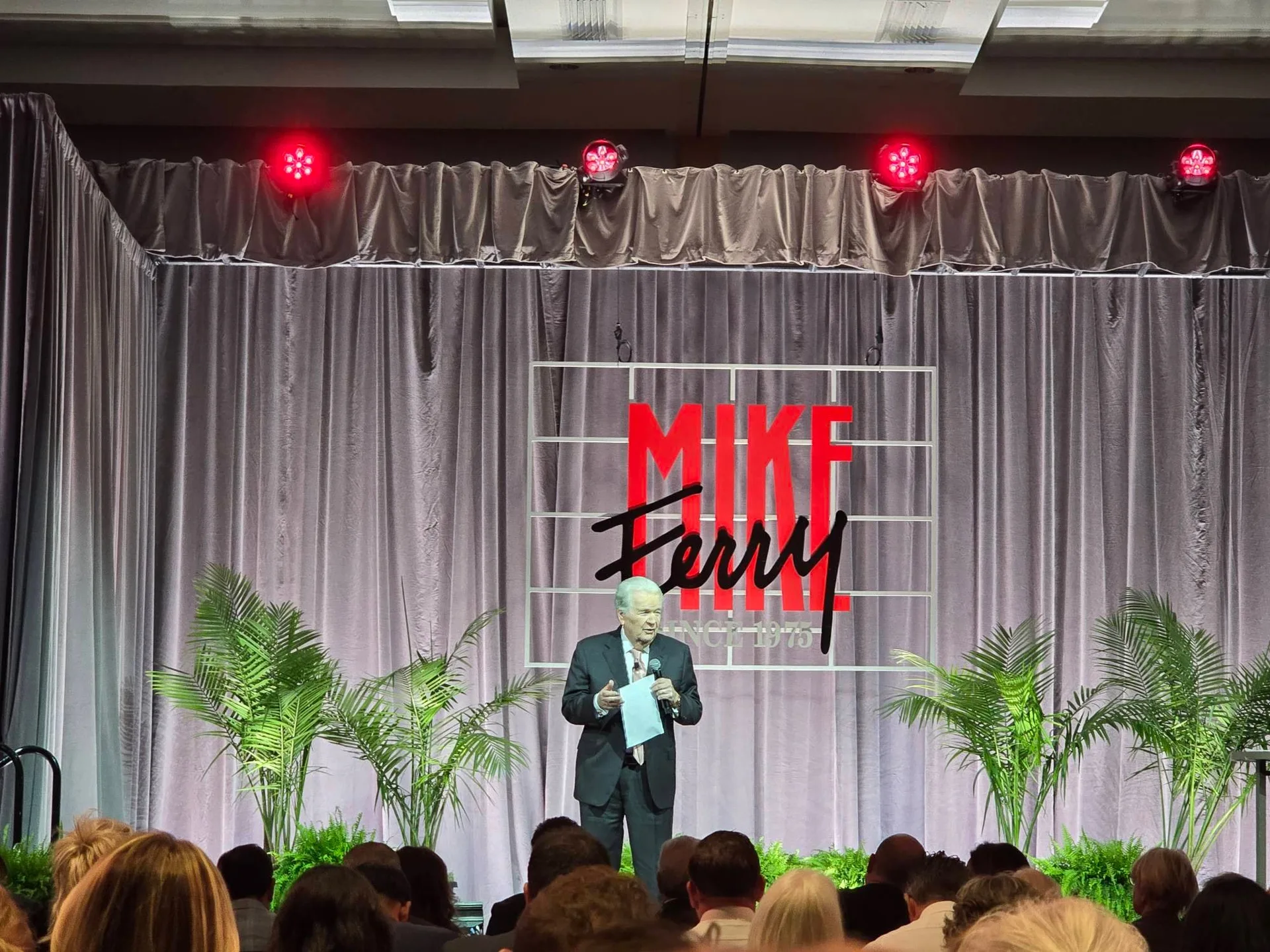 Mike Ferry himself on stage at the podium getting the audience excited at the start of Day 3 at the Mike Ferry 2026 Superstar Retreat at The Diplomat Beach Resort in Hollywood, Florida, with the MIKE FERRY SINCE 1975 backdrop, red stage lights, palm plants, and the audience seated in front