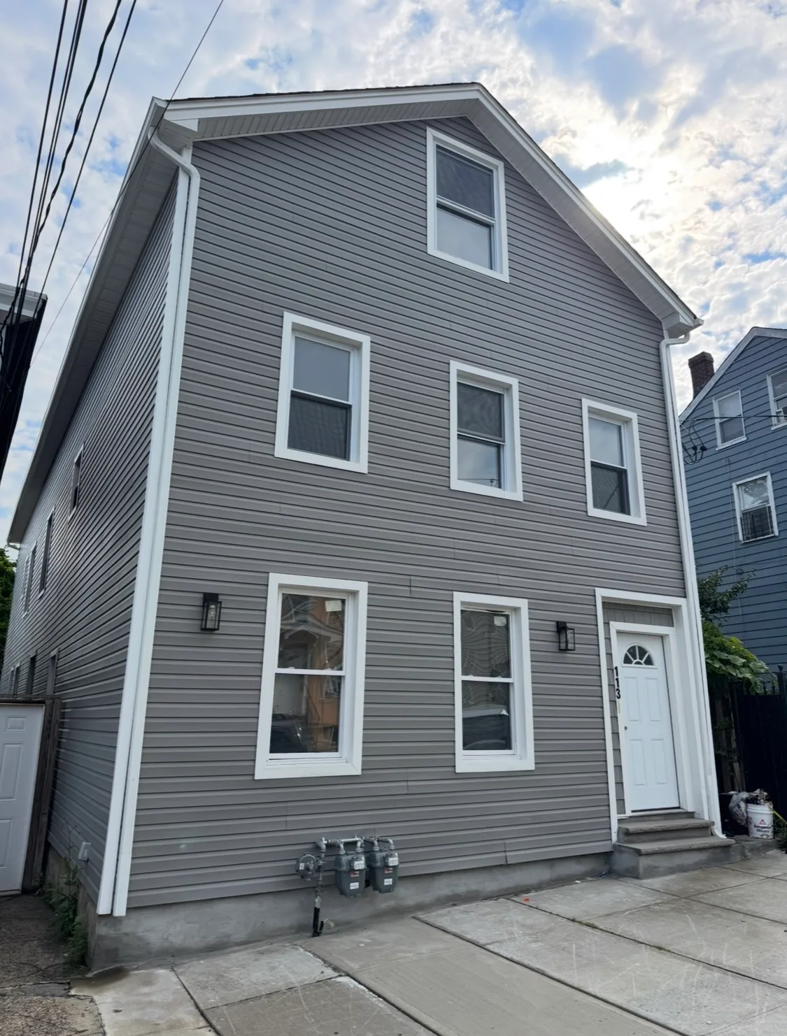 Vertical exterior facade of the three-story building with gray vinyl siding, white trim, front door, and steps.
