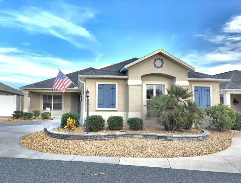 A single-story beige Florida home with blue shutters, a curved stone garden border, and an American flag
