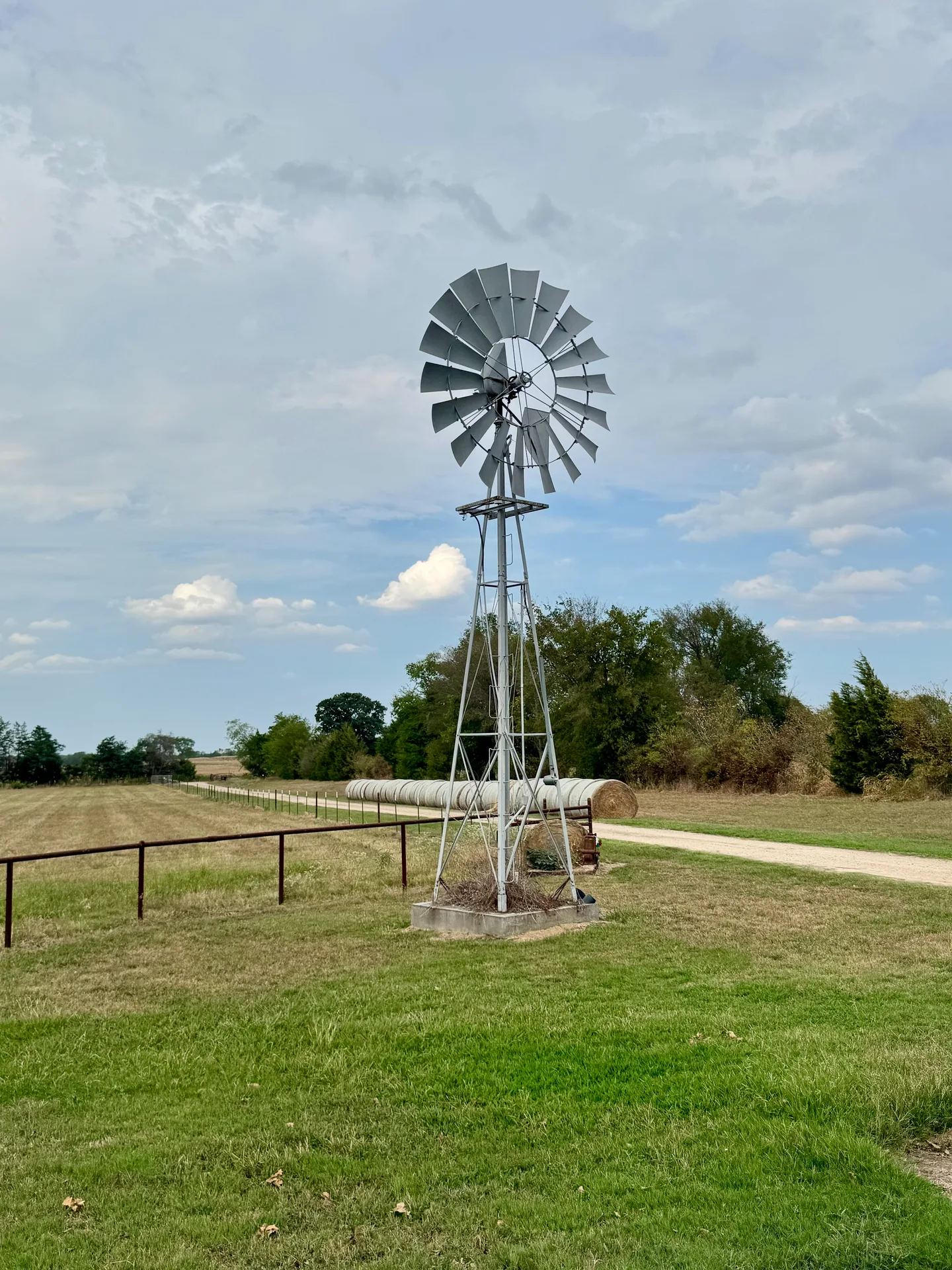 A metal windmill standing in a grassy rural field with hay bales under a cloudy sky in Northeast Texas