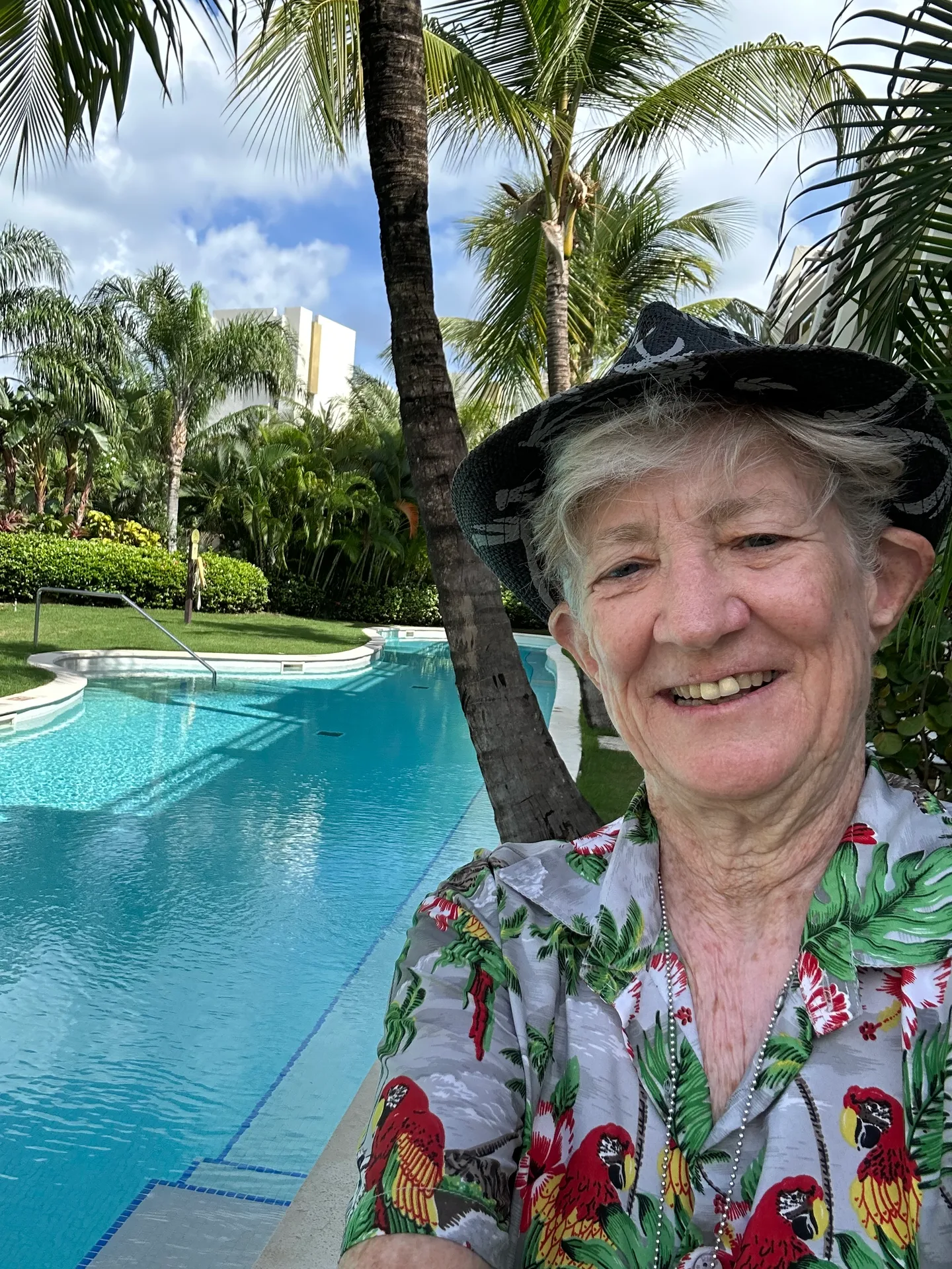 Cynthia Tant, a woman with short grey hair and a warm smile, wearing a tropical shirt and sun hat by a pool.