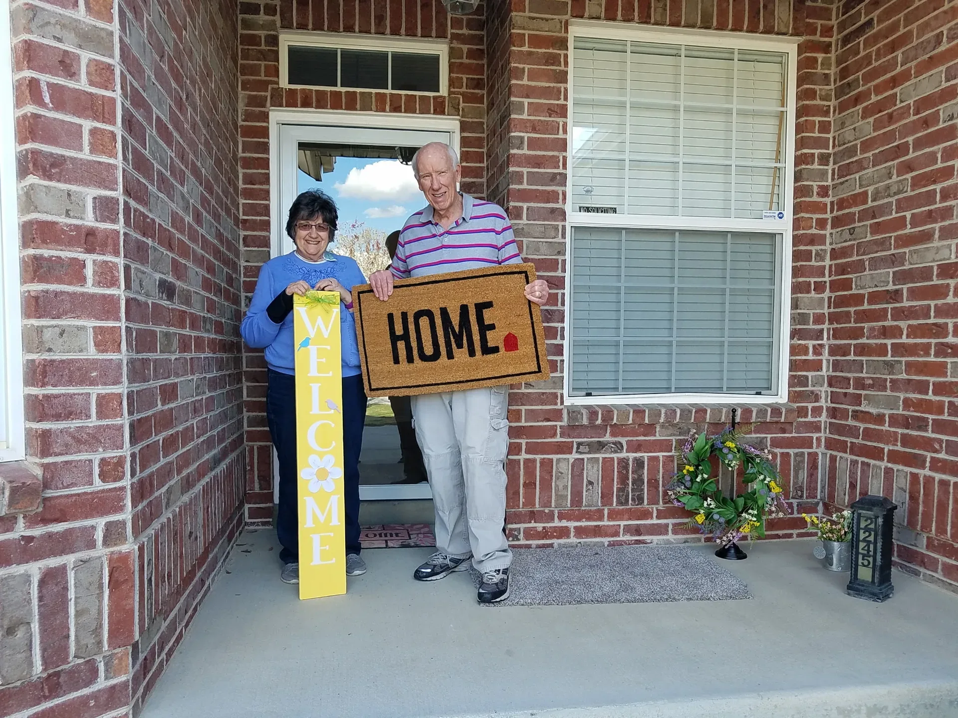 An older couple smiling at the front door of their brick home, the woman holding a tall yellow WELCOME sign and the man holding a HOME doormat