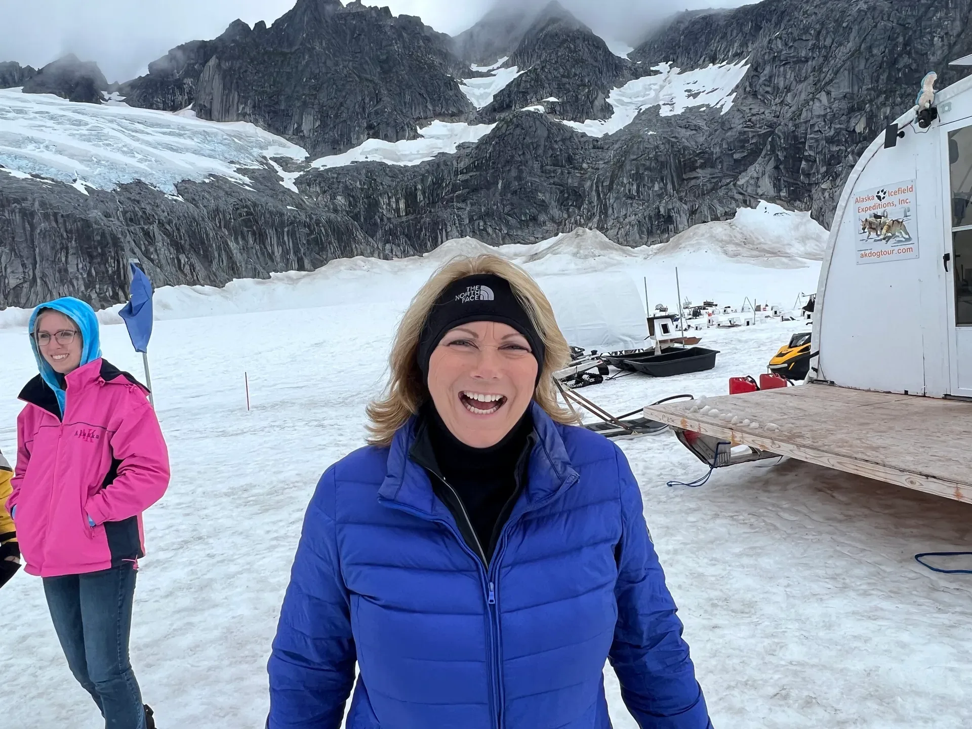 Bev Prock smiling in front of a snow-covered mountain glacier next to a small expedition hut