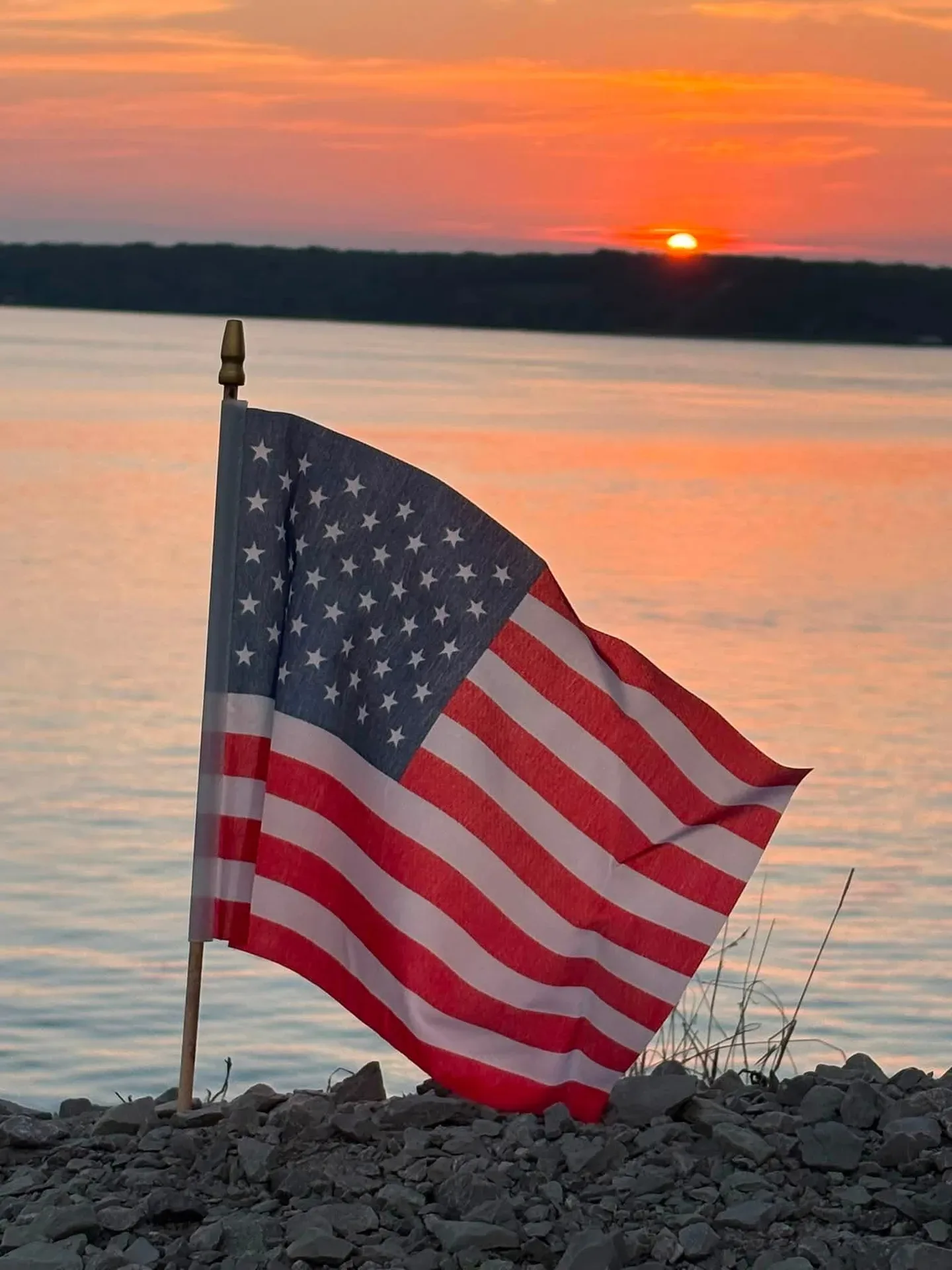 Small American flag planted on rocky ground at sunset over a body of water