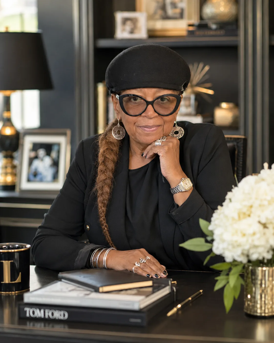 Toni Patillo seated at a desk in a dark, elegant office, wearing a black blazer, beret, and glasses
