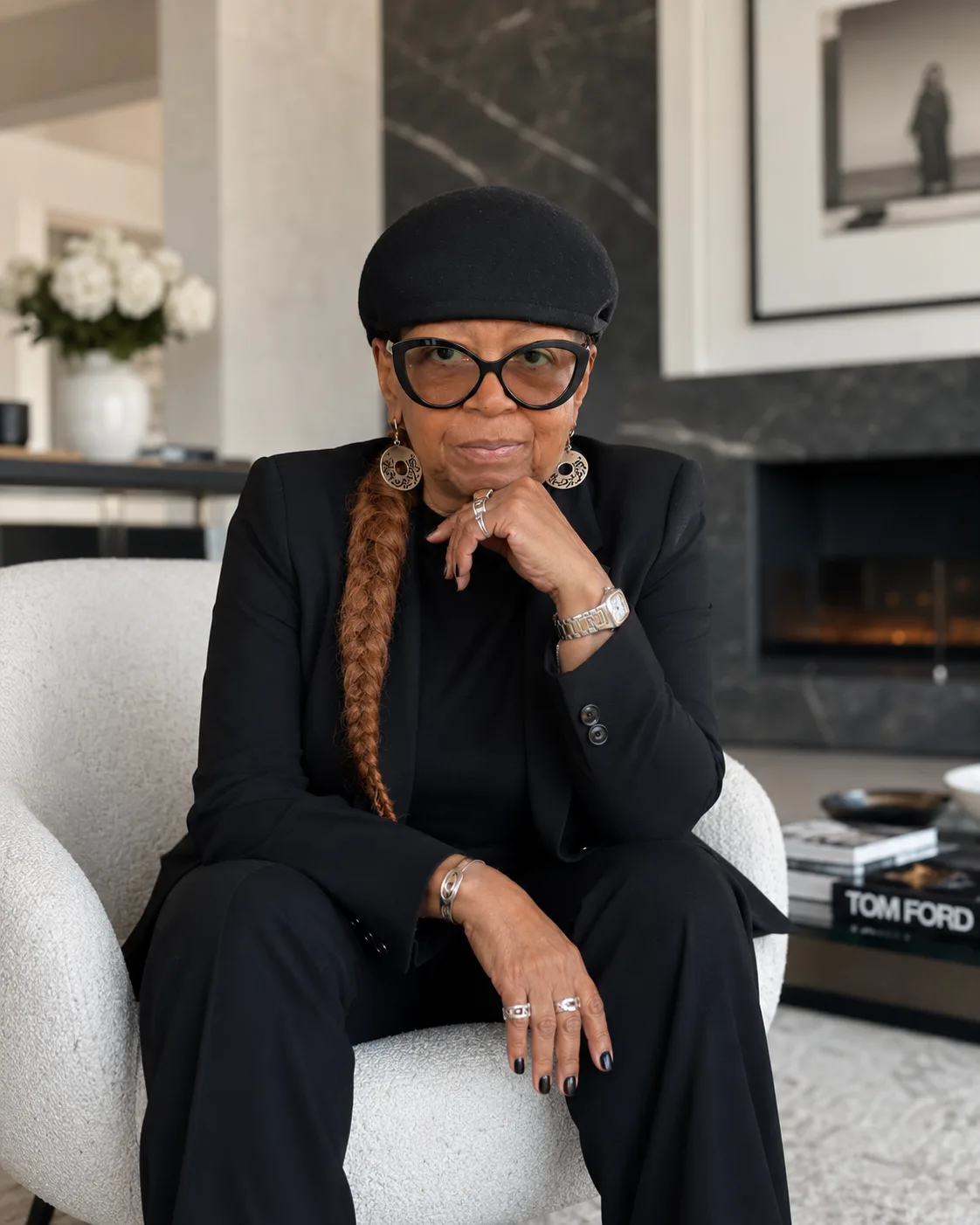 Toni Patillo, founder of Journey Beyond Fifty, seated in a white armchair in a modern living room, wearing a black blazer, hat, and glasses