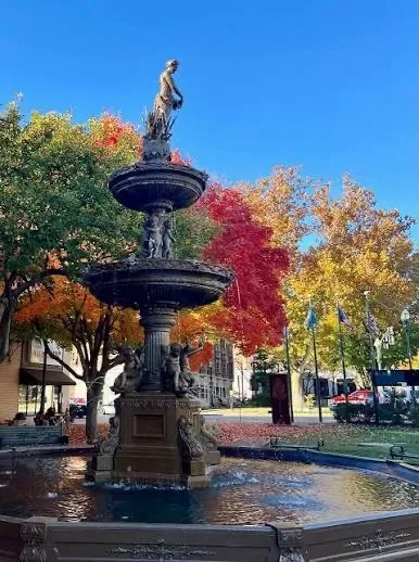 A large tiered outdoor fountain surrounded by mature trees in full autumn color under a clear blue sky, the kind of public green space that anchors park-adjacent neighborhoods across Greater Cleveland