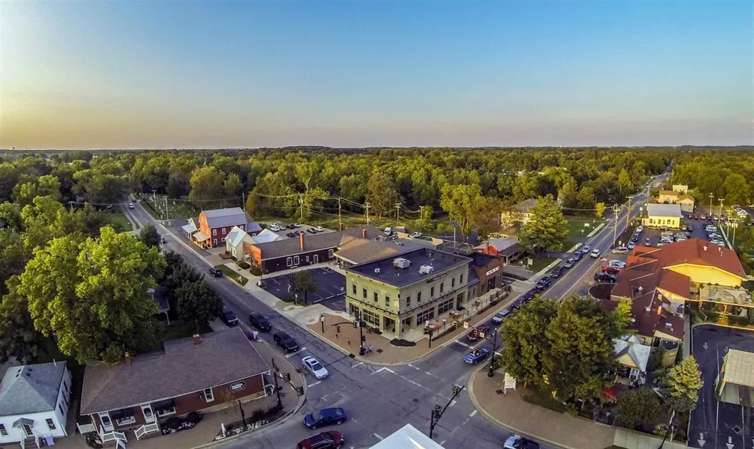 An aerial view of a small Ohio town street corner with mixed-use buildings and shops surrounded by mature trees on a clear autumn afternoon, warm light across the rooftops