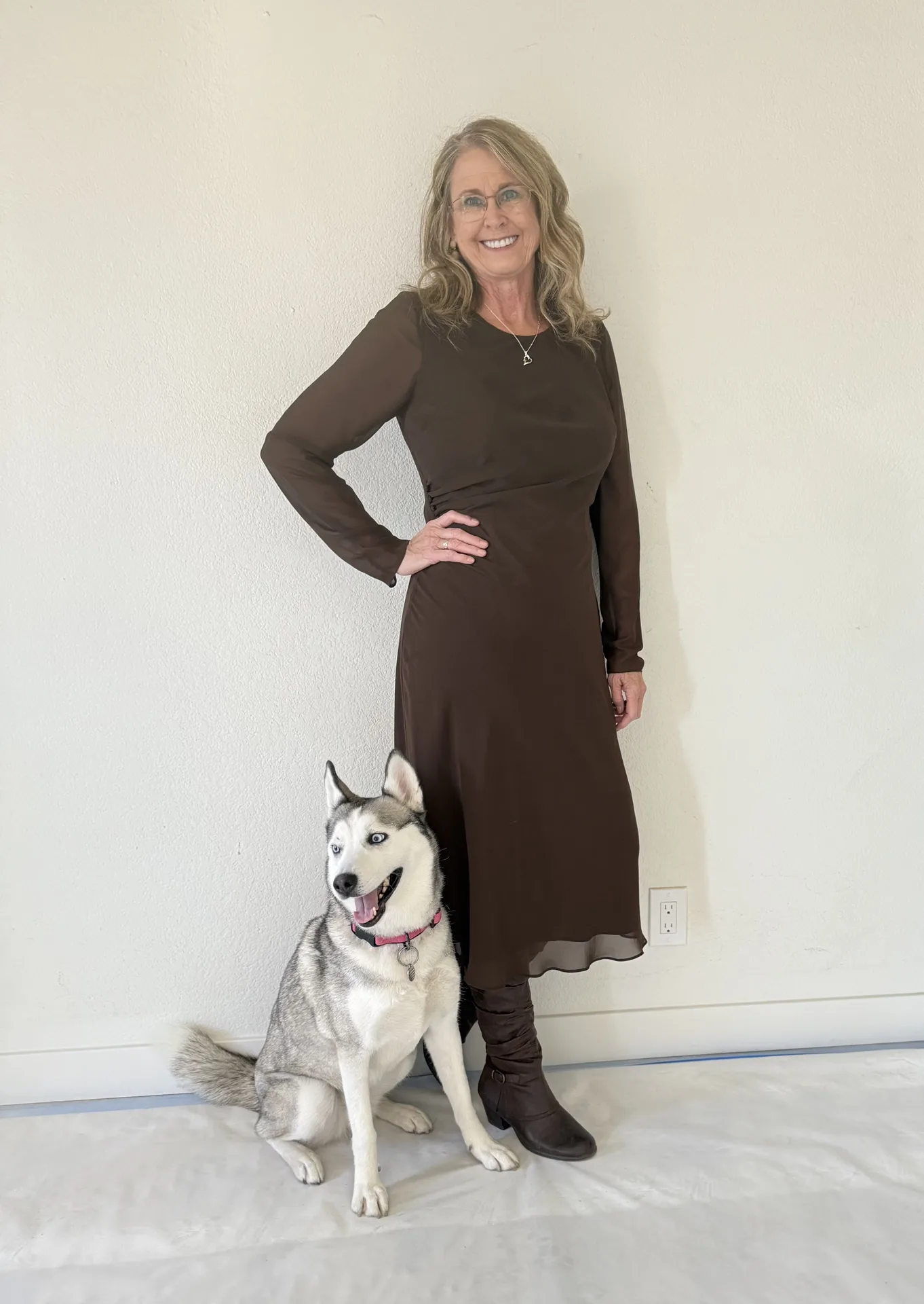 PJ Selby, a smiling woman with wavy blonde hair in a brown dress, standing beside a husky