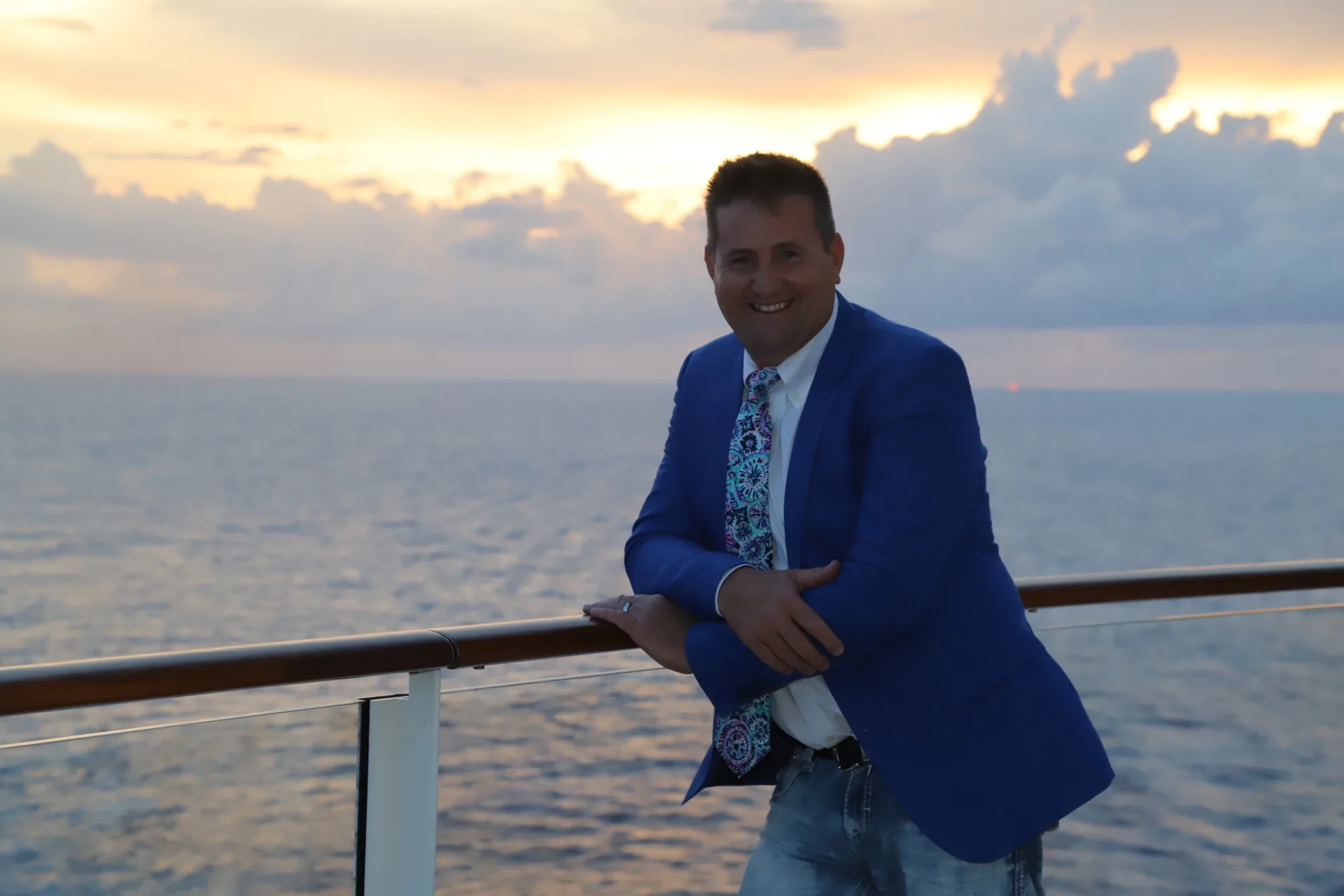 Rob Dietrich in a blue blazer leaning against a cruise ship railing at sunset